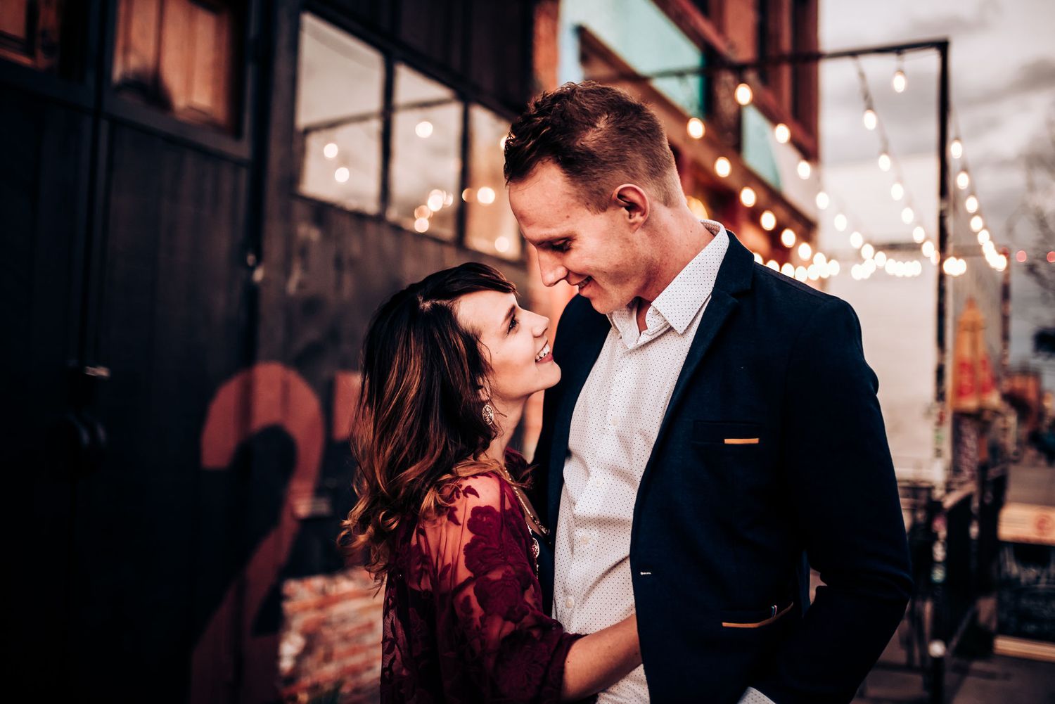 Couple shares tender moment against industrial building backdrop at dusk.