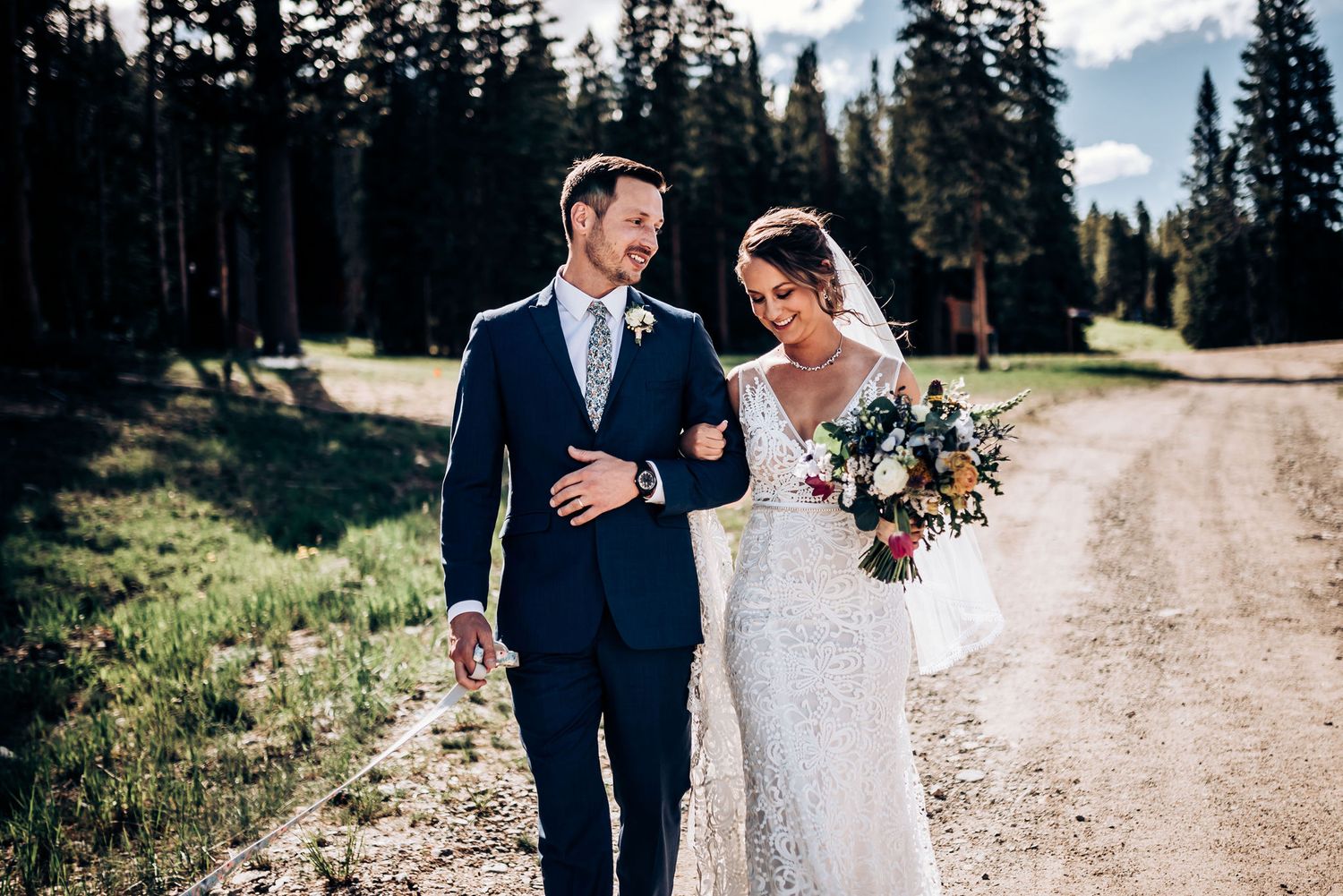A wedding couple walks down a mountain dirt road surrounded by pine trees.