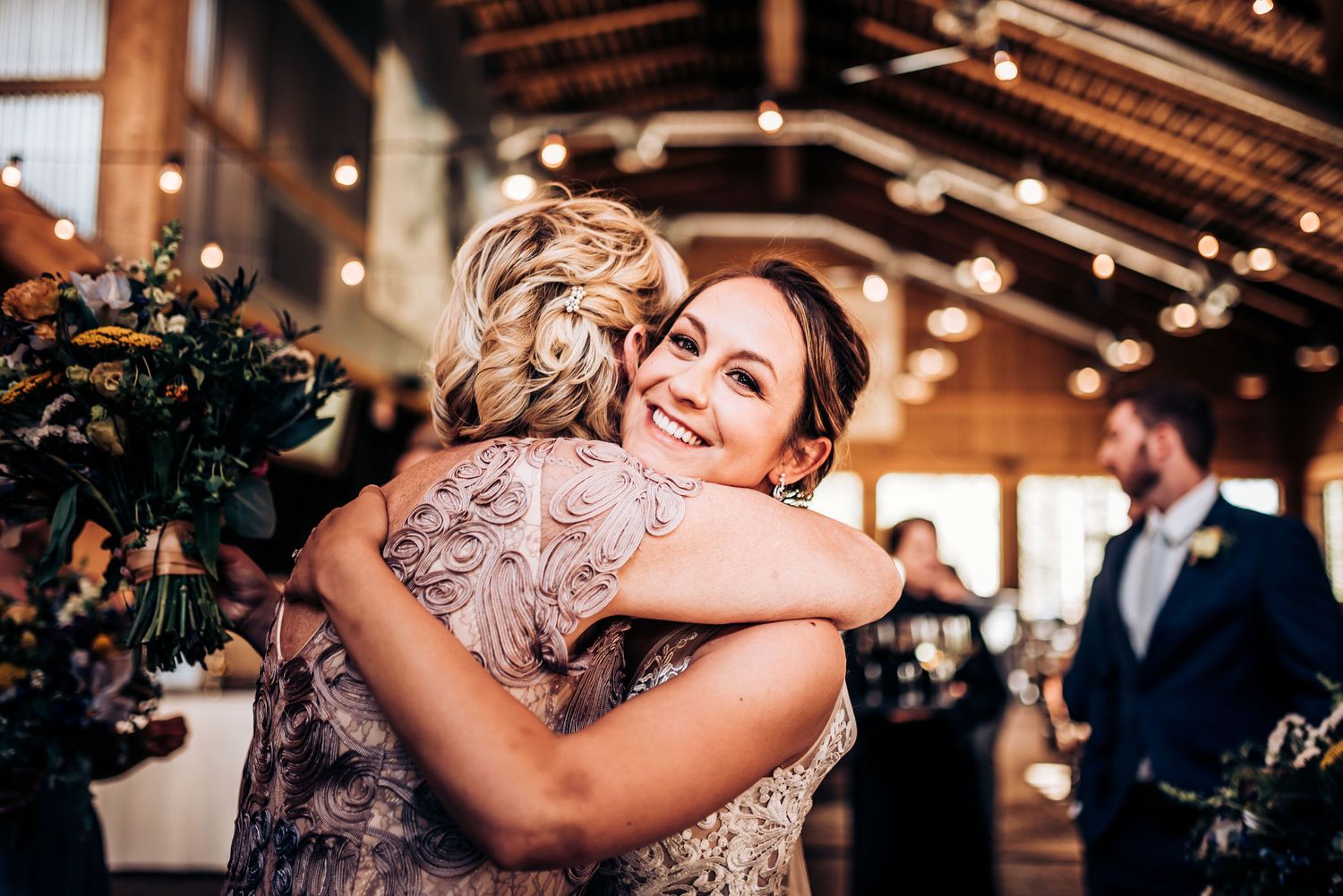 Warm embrace shared during reception in rustic barn venue with string lights overhead.