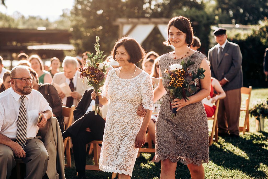 Wedding attendees walk down an outdoor aisle carrying floral bouquets in warm sunlight.