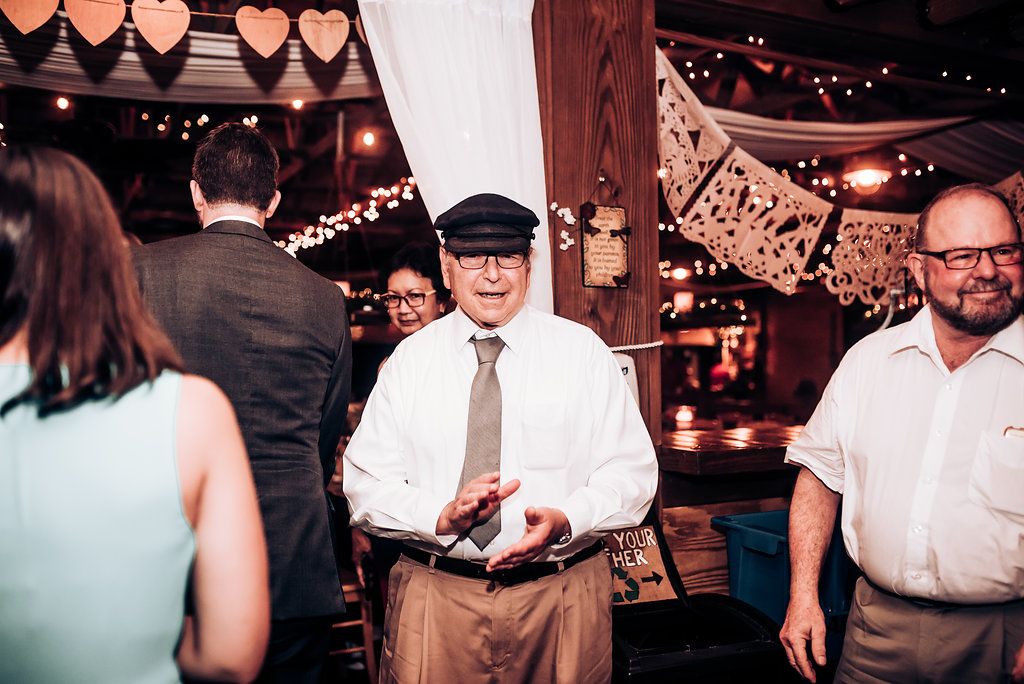 Wedding reception guests dance and mingle under decorative string lights at an indoor venue.
