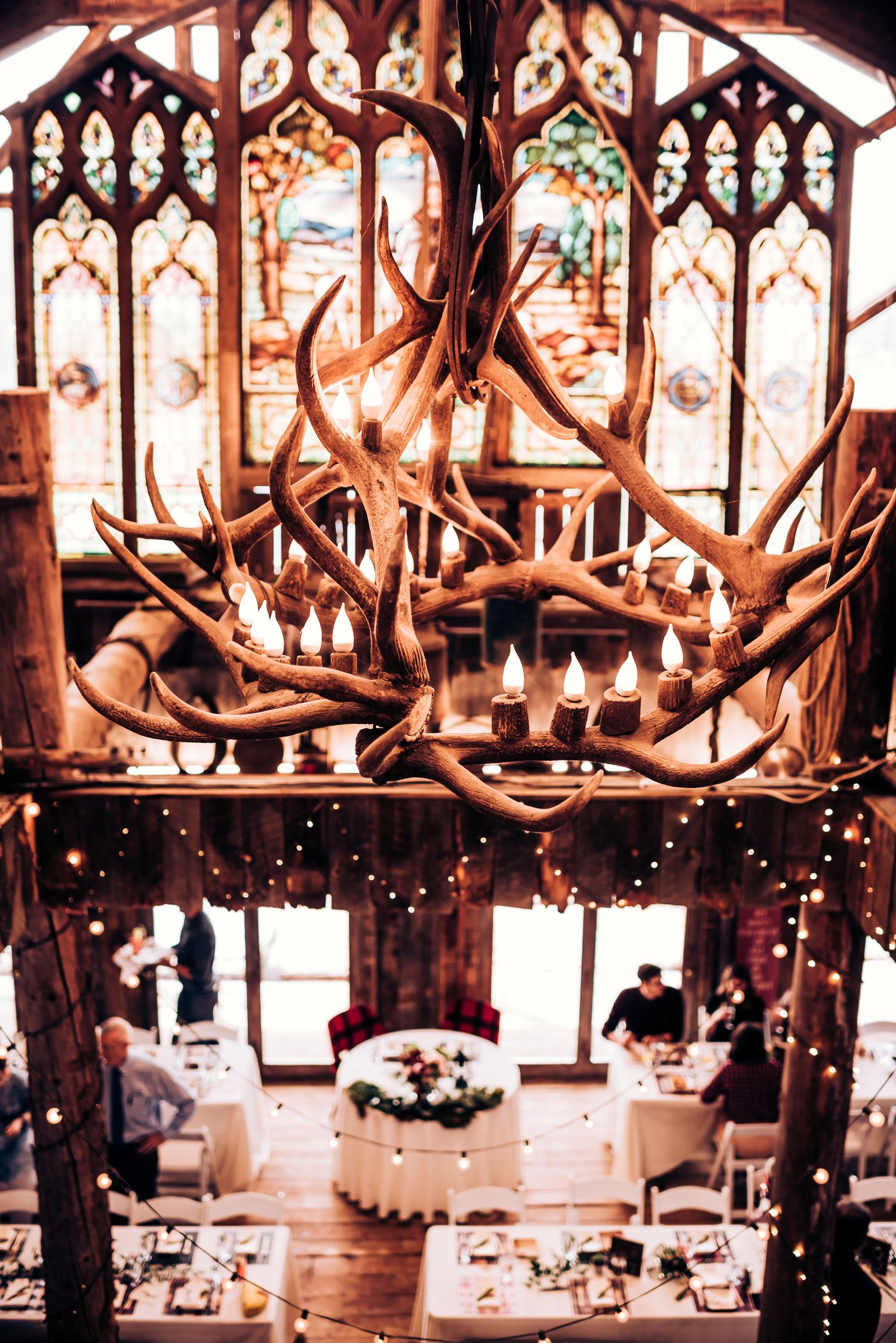 A rustic wedding reception venue with antler chandeliers and gothic windows illuminated by string lights.