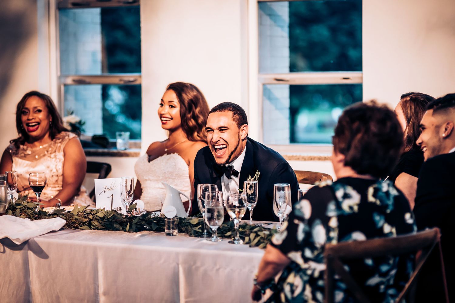 Elegant wedding reception at head table with guests enjoying dinner and laughter.