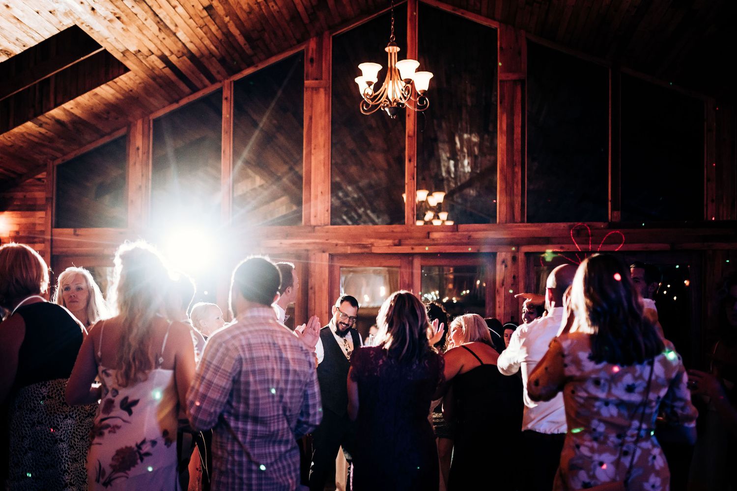 Guests dancing under wooden beams and chandeliers at rustic indoor venue.