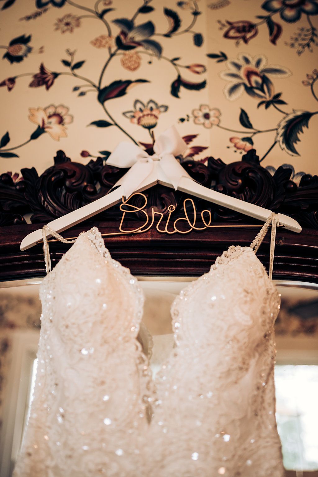 White lace wedding dress on custom bride hanger against floral wallpaper background.