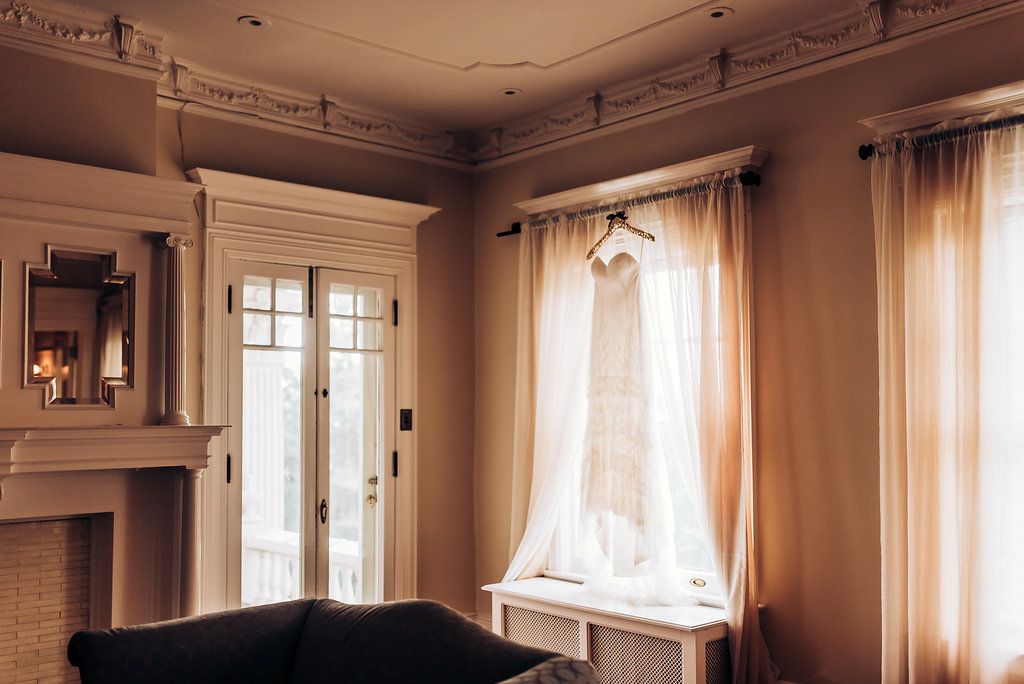 A delicate white wedding dress hangs by a sunny window in an elegant room with ornate molding.