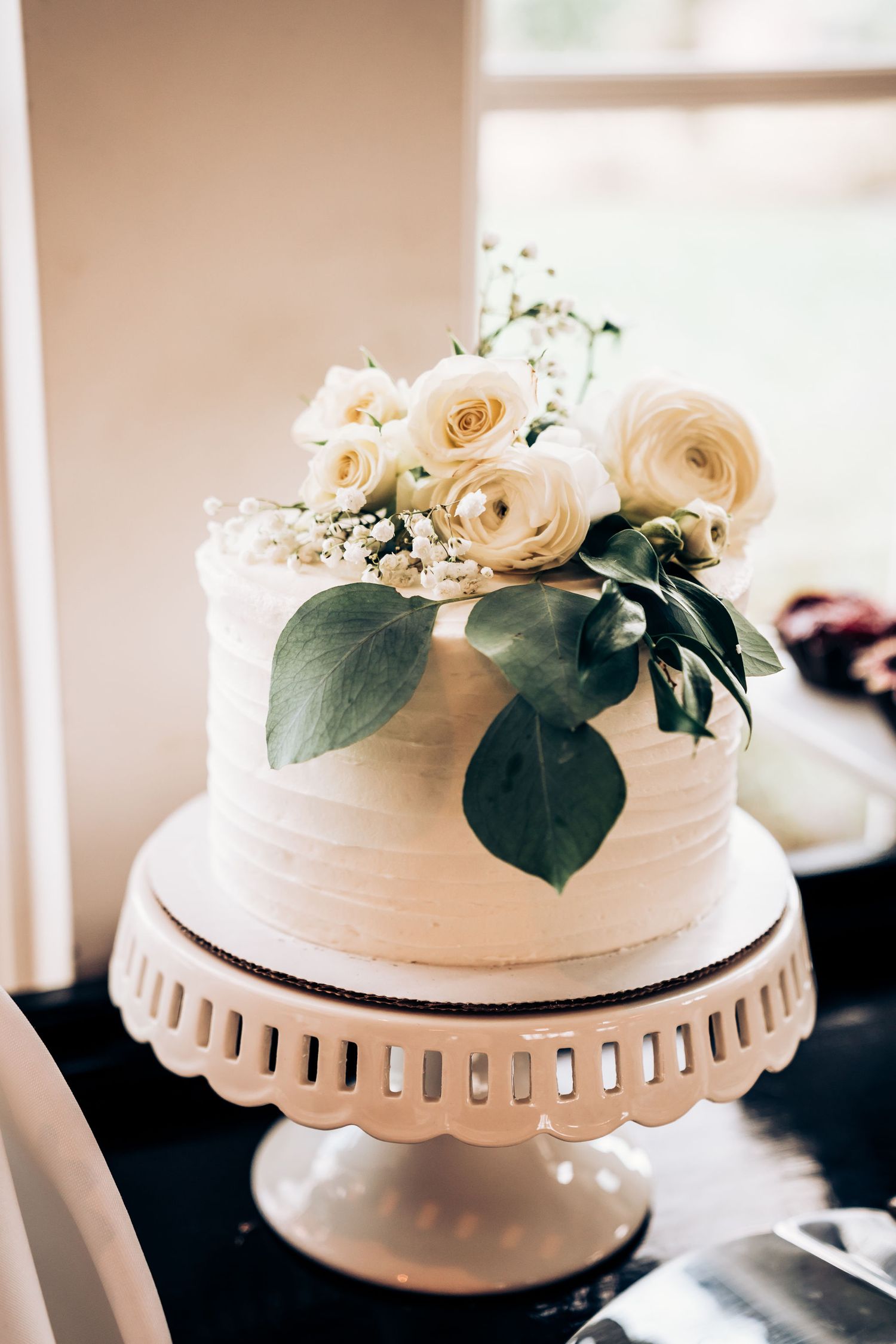 White wedding cake decorated with cream roses and green foliage on a vintage pedestal stand.