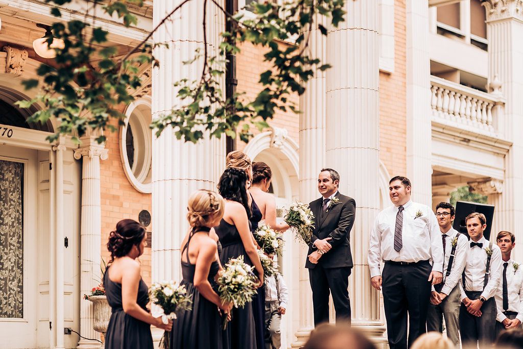 Wedding party in navy dresses and white shirts gather on stone steps during outdoor ceremony.