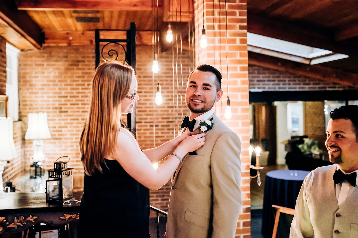 Pre-wedding preparation scene with bow tie adjustment in rustic brick venue.