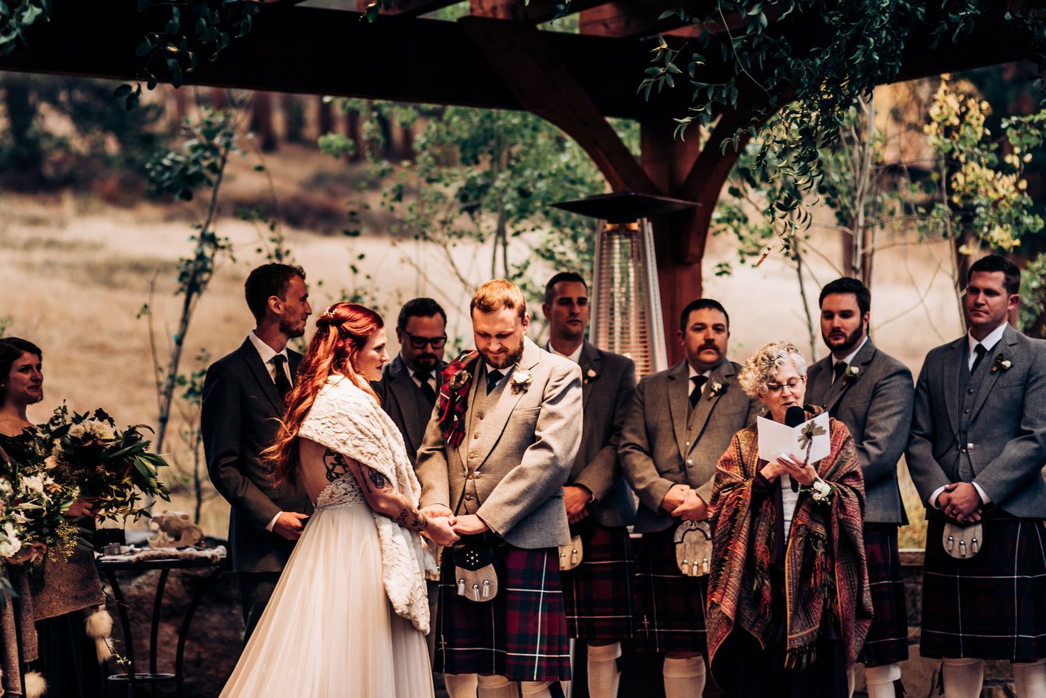 Wedding ceremony under trees with bridal party wearing traditional Scottish kilts and formal attire.