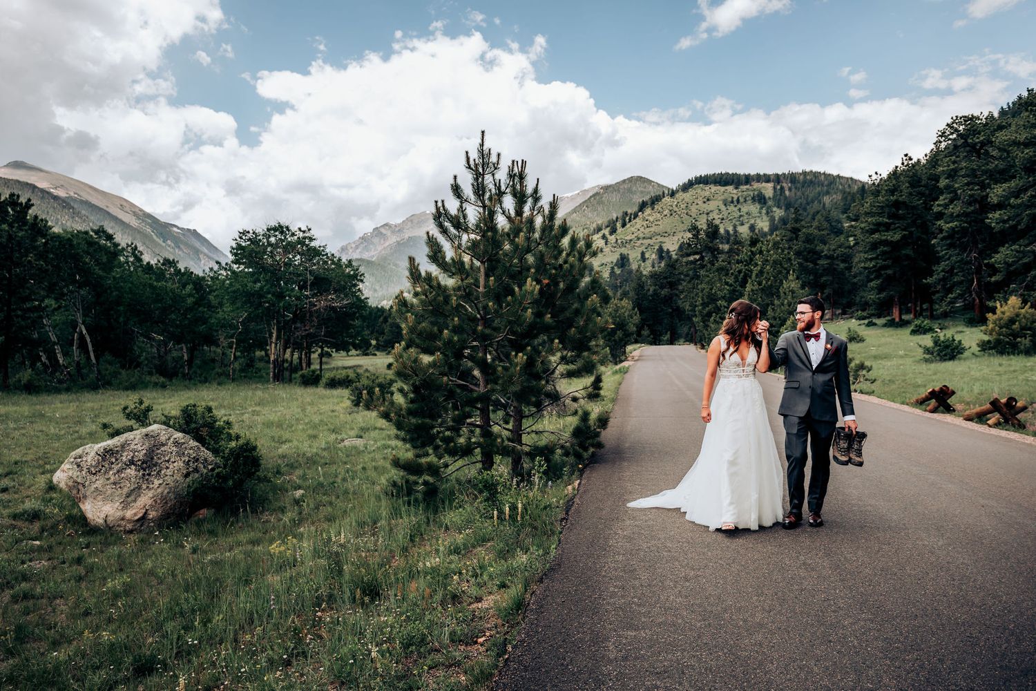 A newlywed couple walks along a scenic mountain path surrounded by evergreen trees and rocky terrain.
