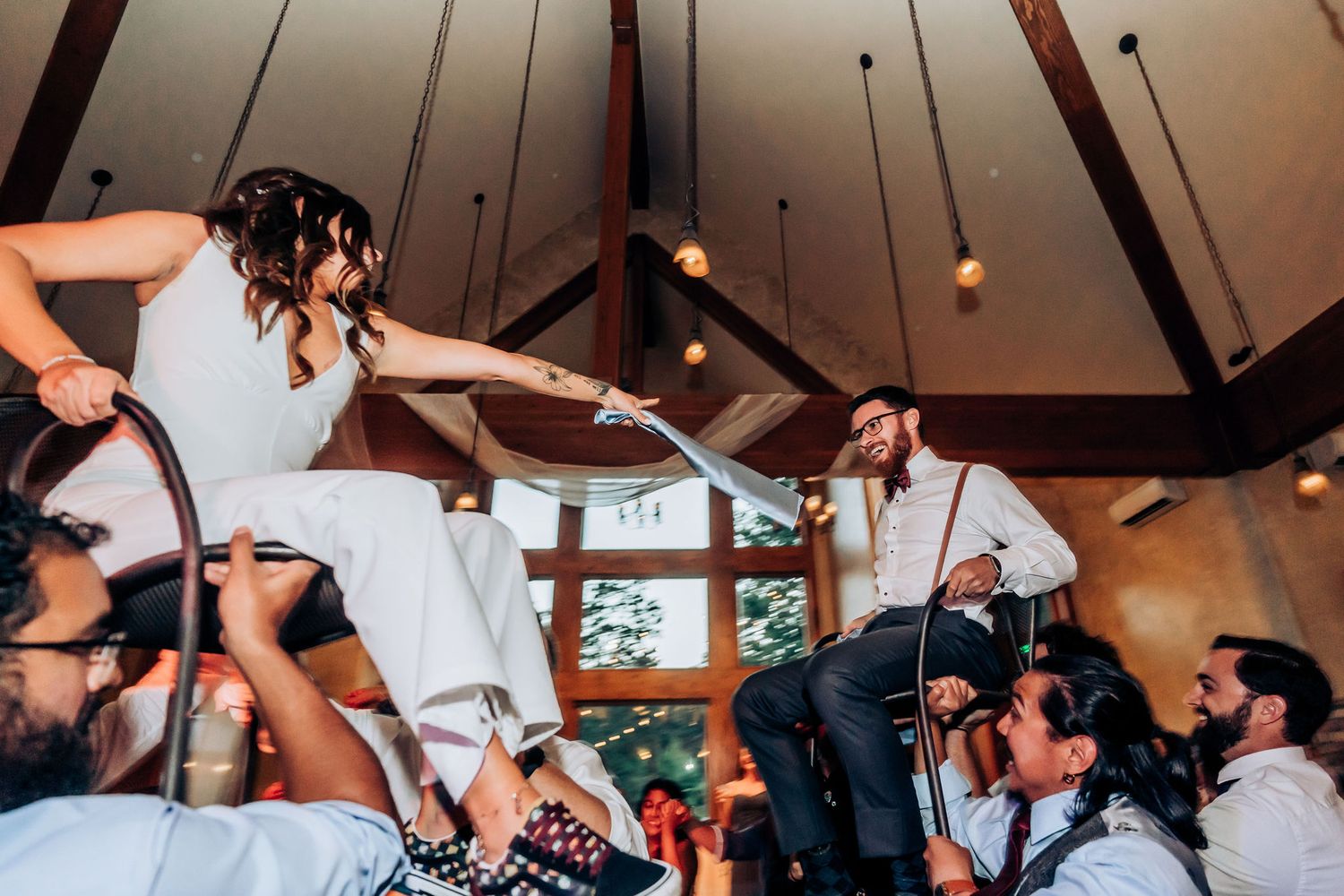 Traditional wedding dance with guests lifting celebrants on shoulders in a rustic venue.