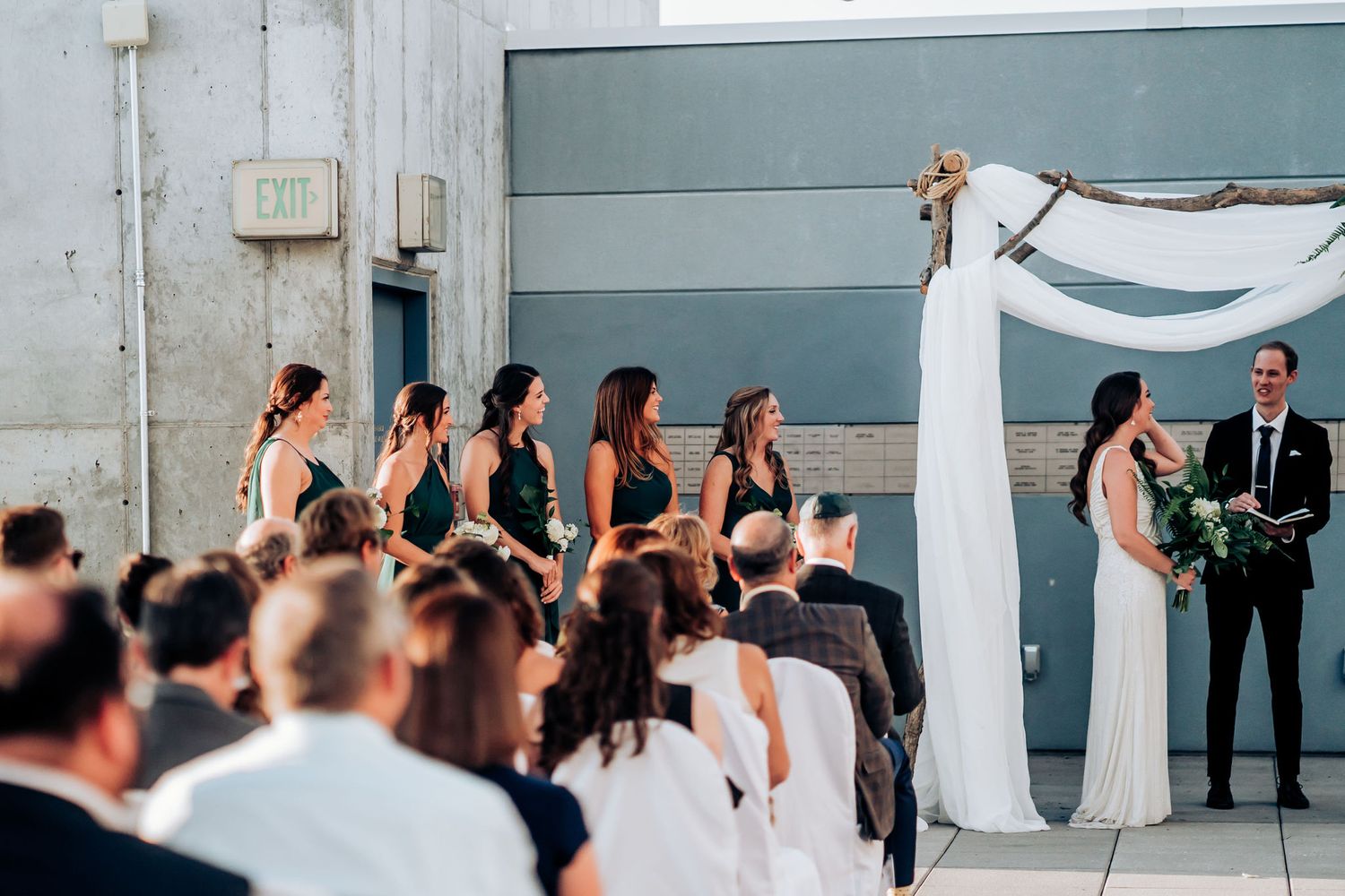 Outdoor wedding ceremony with bridesmaids in black dresses and white-clad guests.