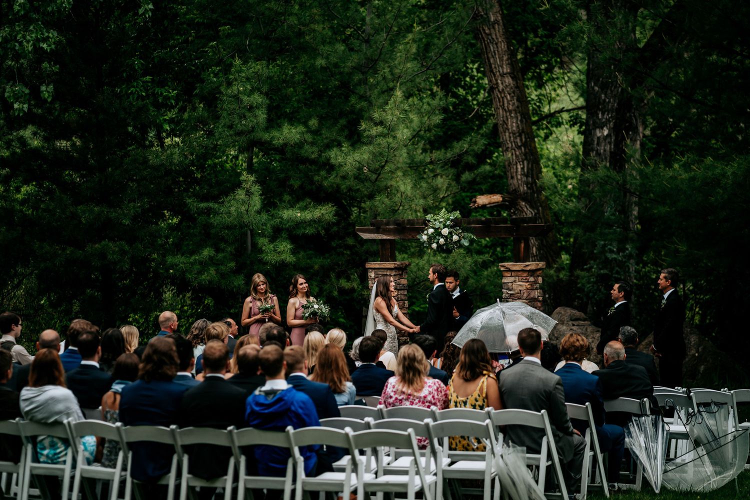 An outdoor wedding ceremony with guests seated in white chairs surrounded by lush forest scenery.