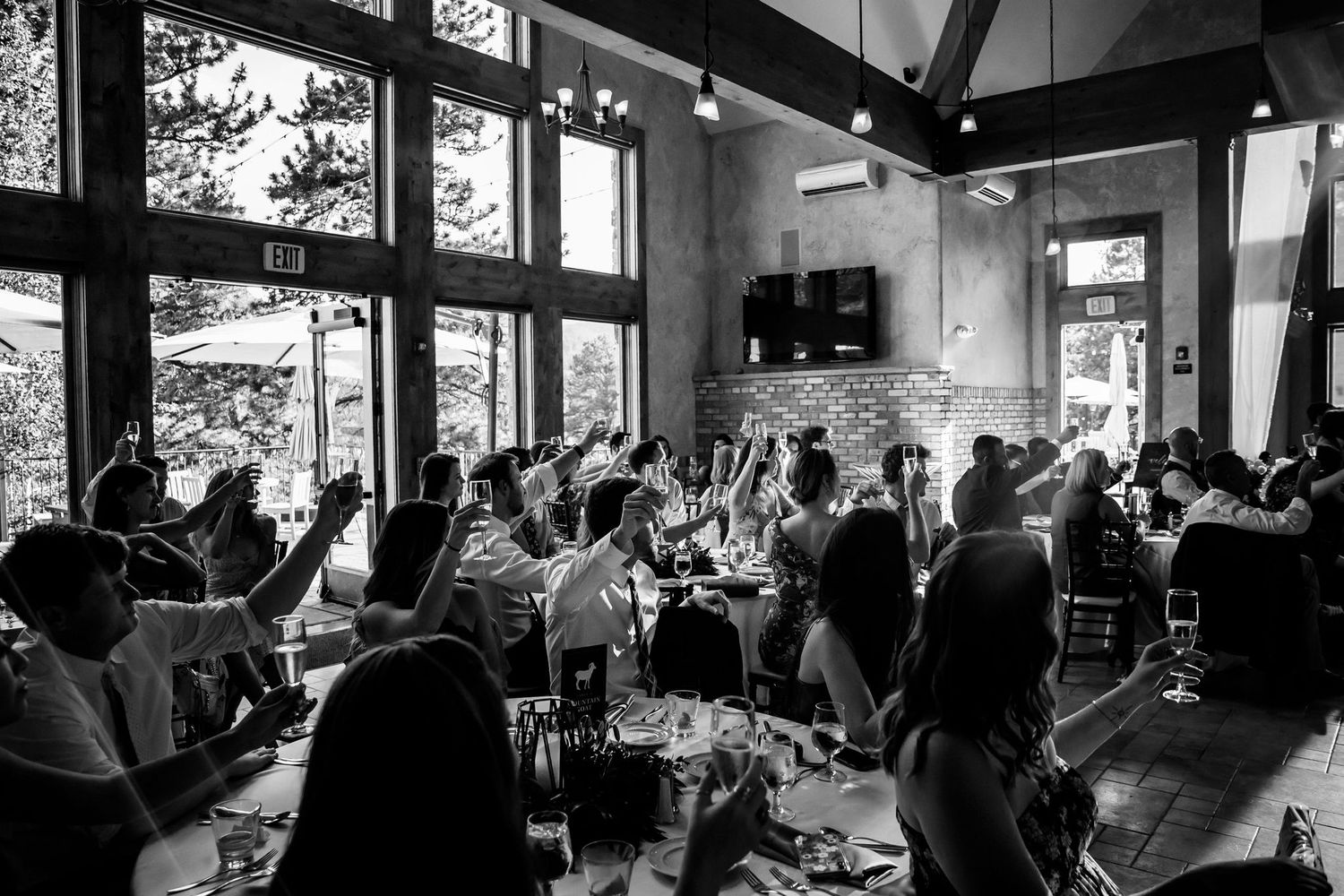 Black and white photo of wedding guests enjoying the reception in a bright, window-lit venue.