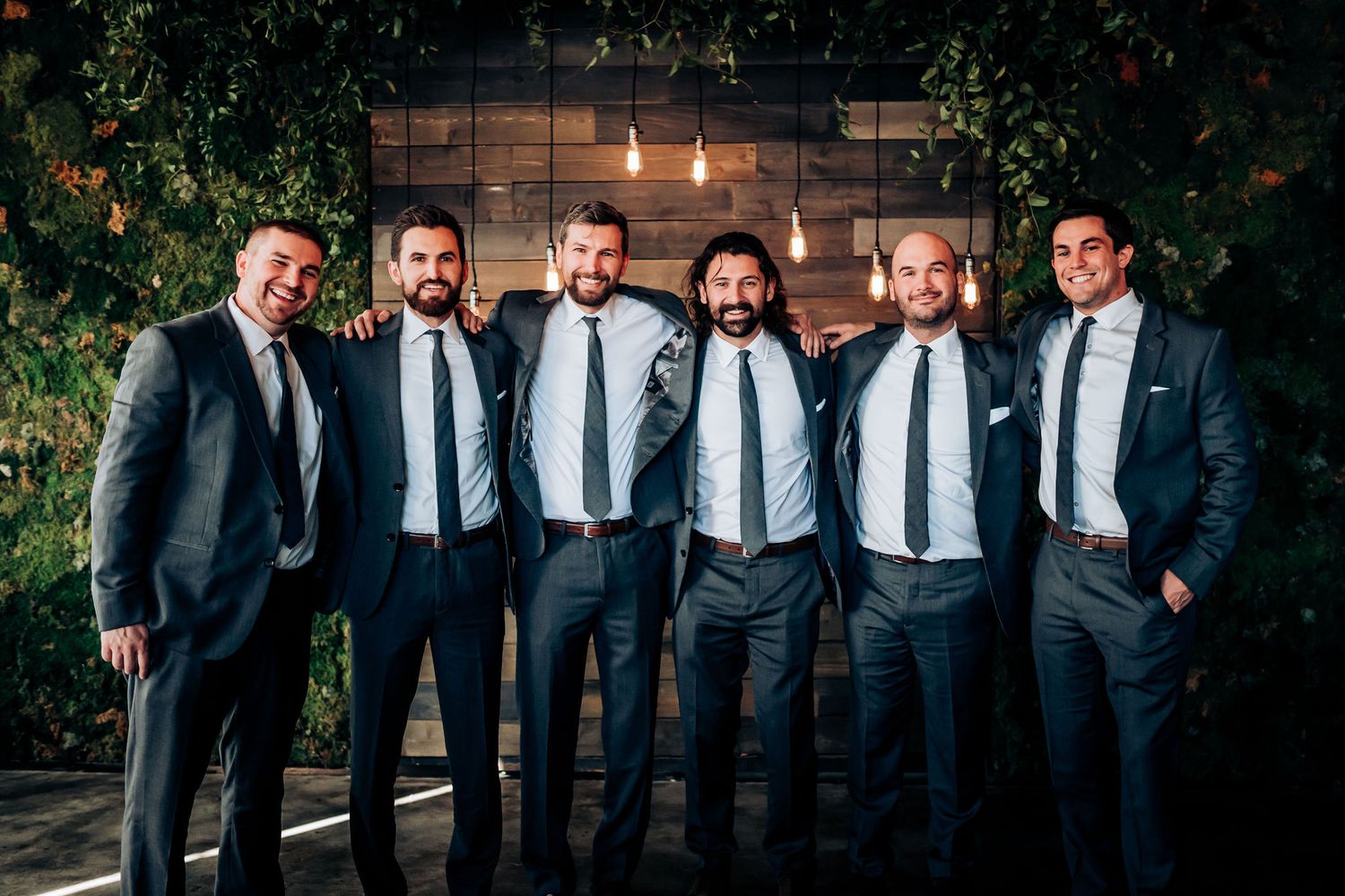 Group of groomsmen in matching dark suits smile together in front of an illuminated wall with foliage.