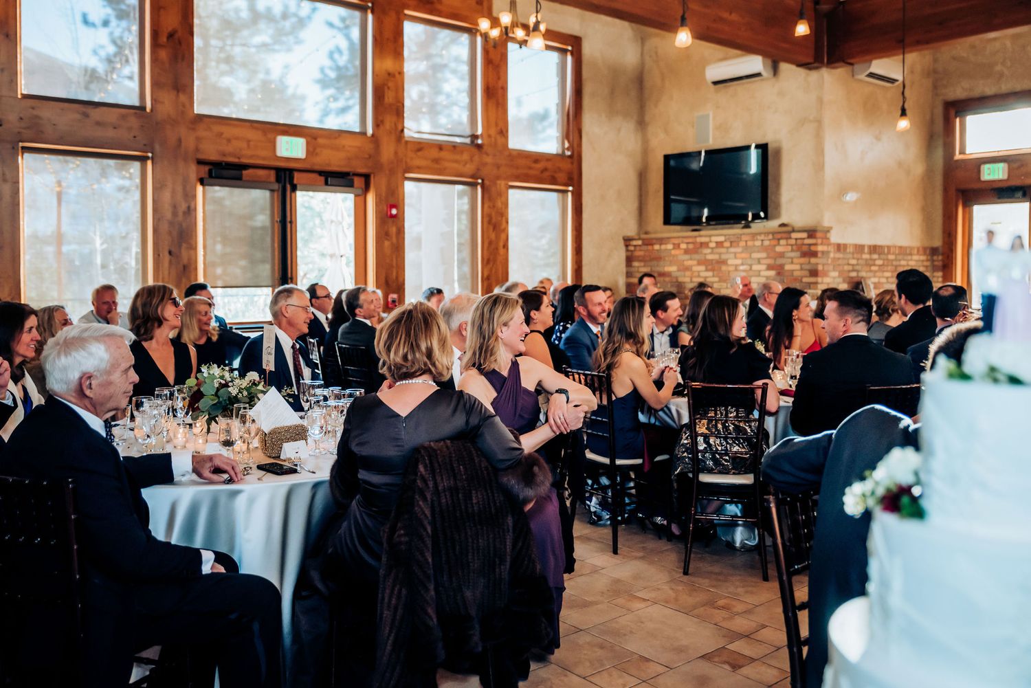 Wedding reception venue filled with guests at round tables in rustic wood-paneled room.
