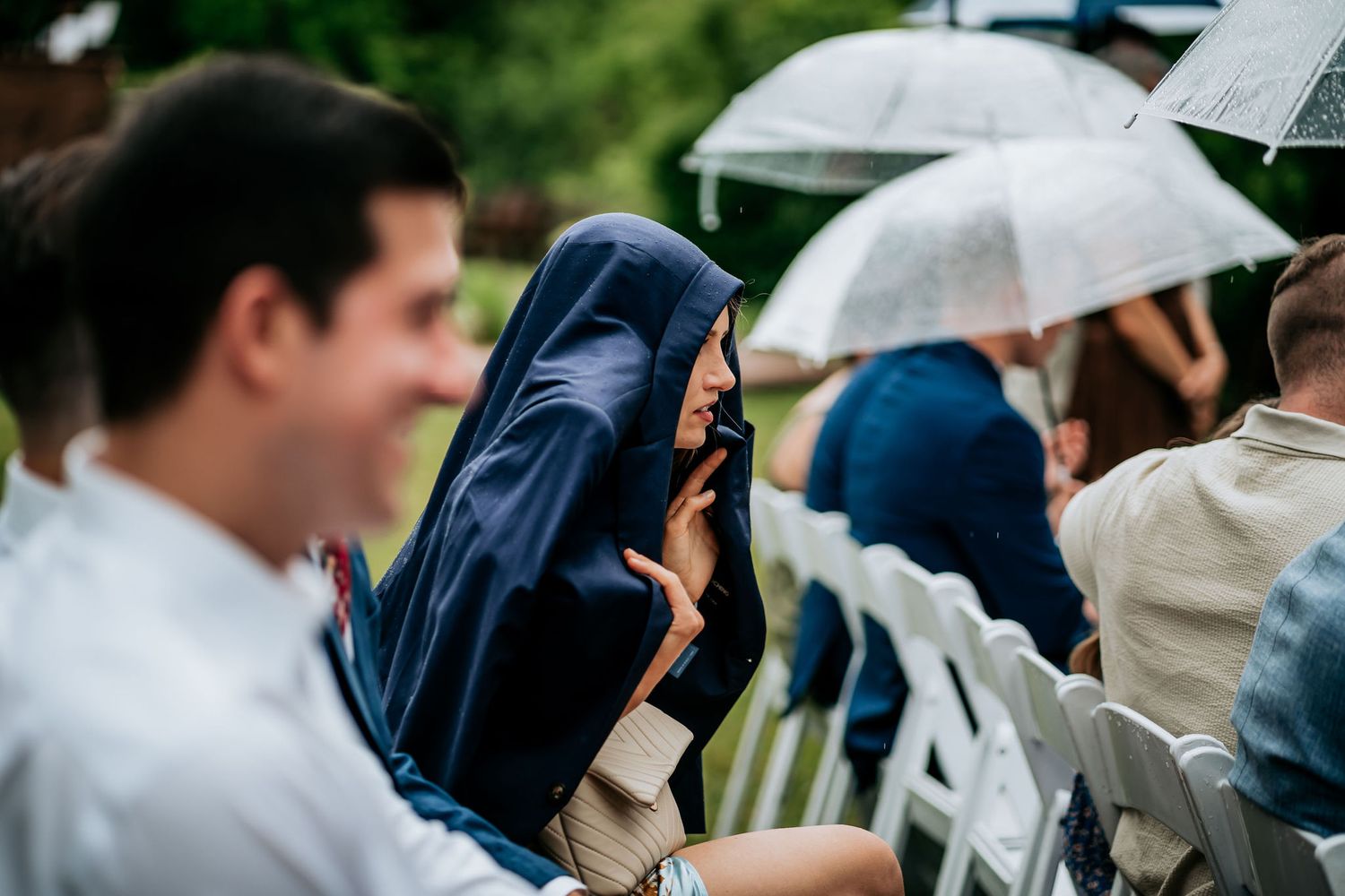 Wedding guests sit in rows of white chairs holding umbrellas during an outdoor ceremony.