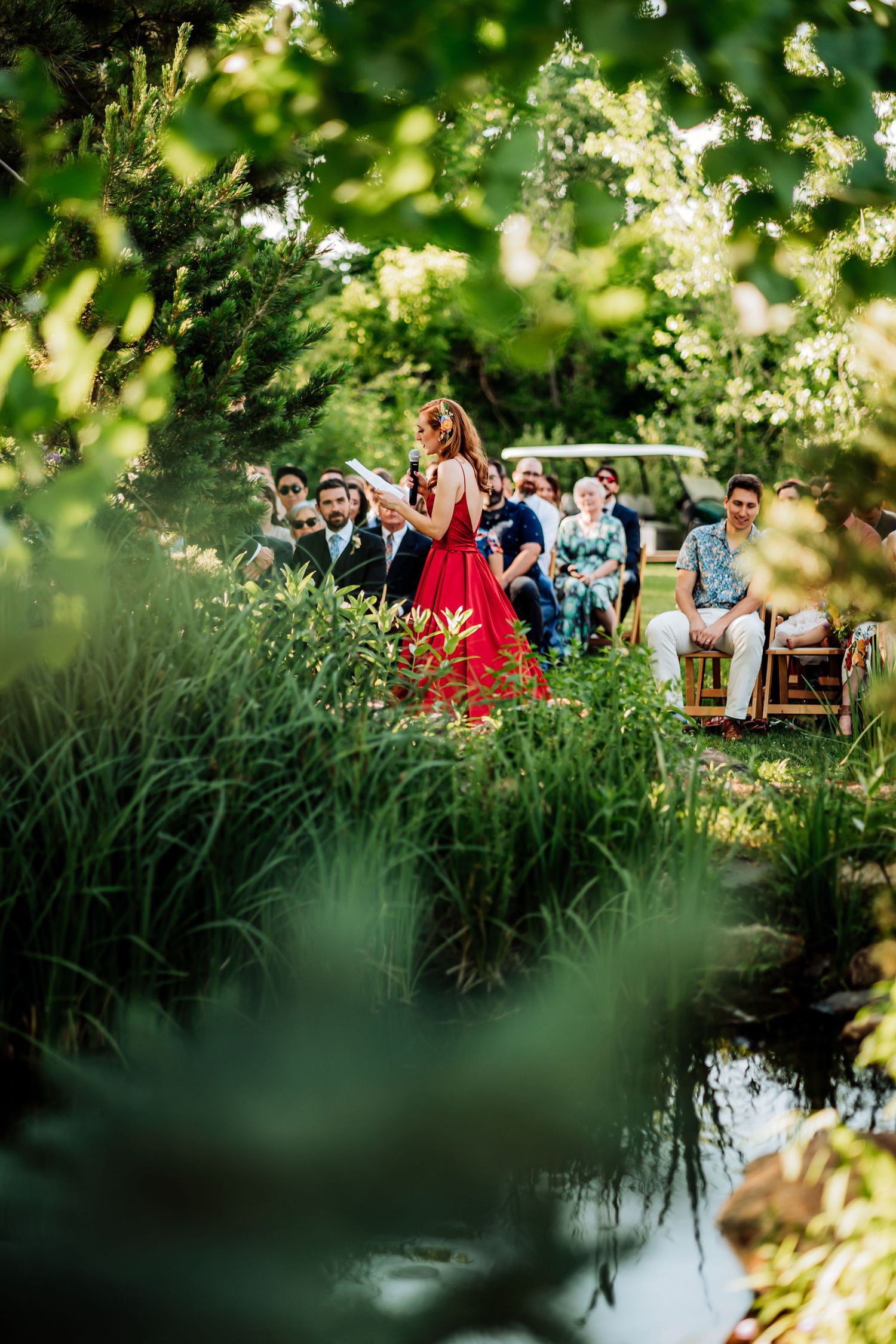 An outdoor wedding ceremony viewed through greenery with guests seated by a pond.