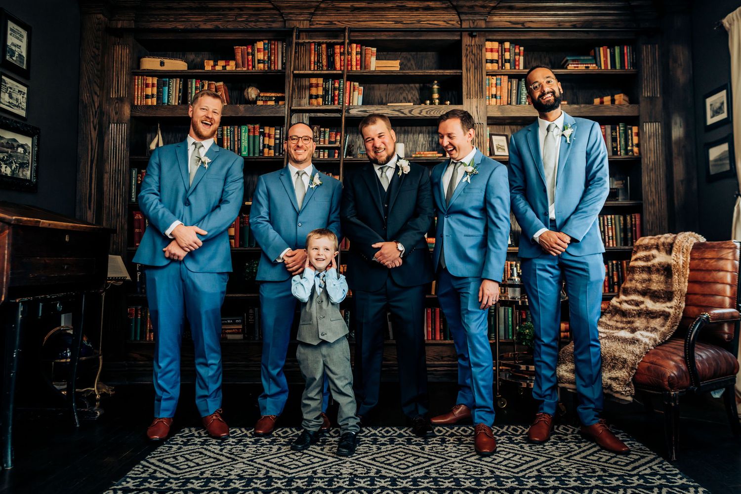 Wedding party in matching blue suits poses in elegant wood-paneled library with bookshelves.