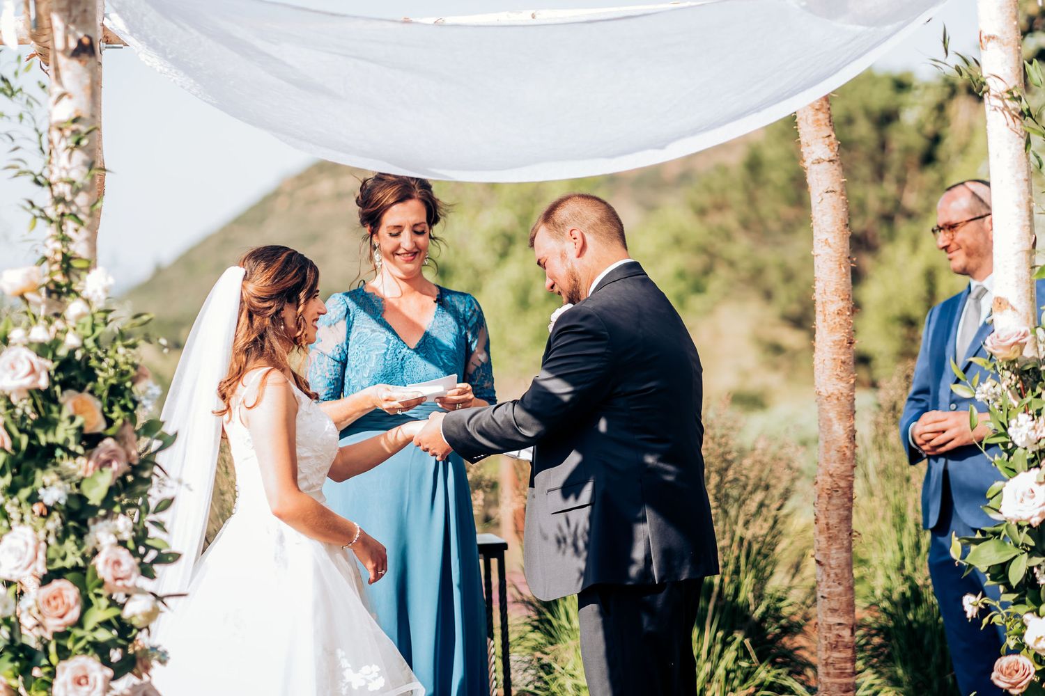 Wedding ceremony exchange of rings under white-draped wooden arch with floral accents.