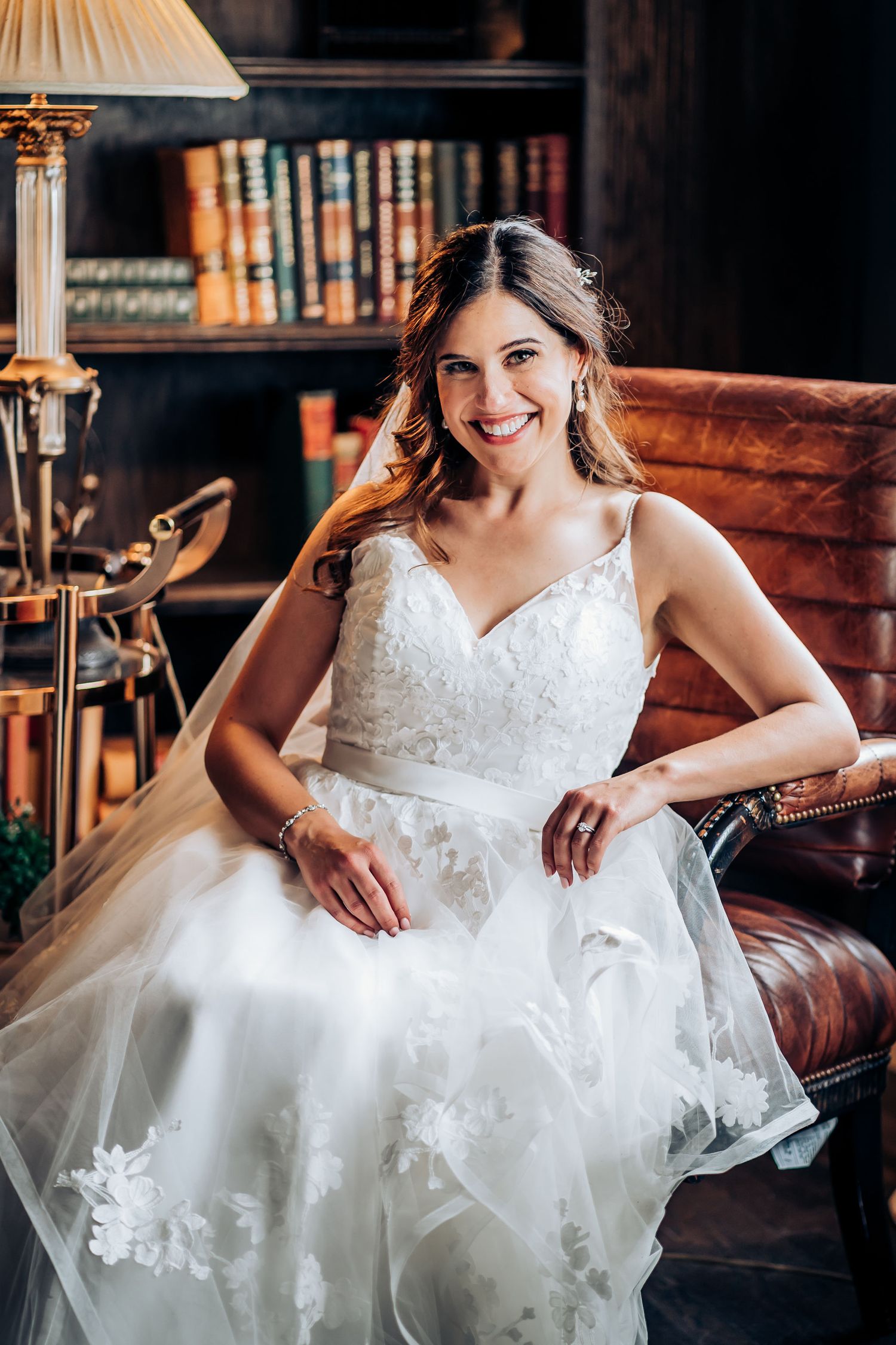 Bridal portrait in white lace gown seated in leather chair with library bookshelf background.