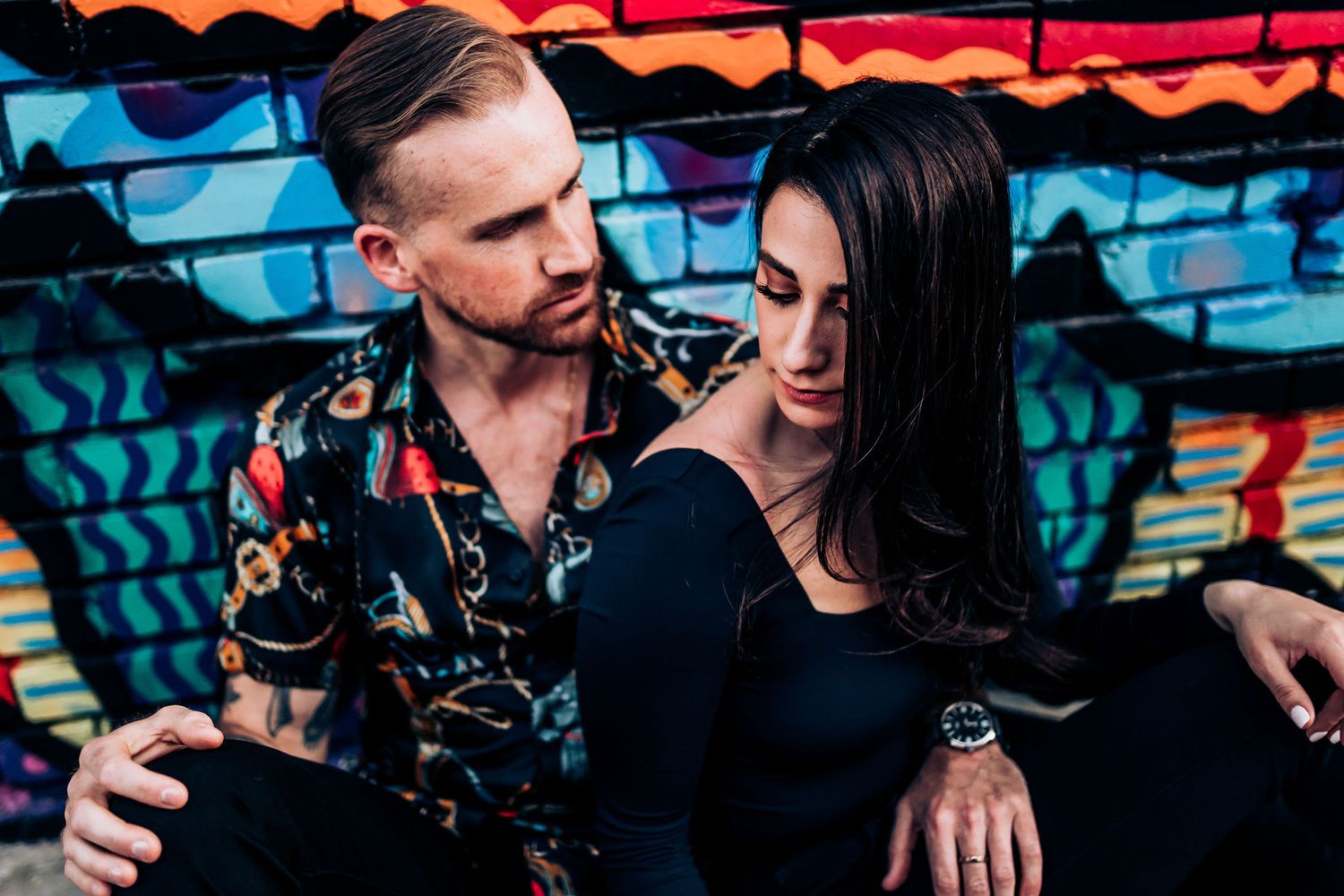 A couple poses intimately against street art while one wears a patterned Hawaiian shirt.