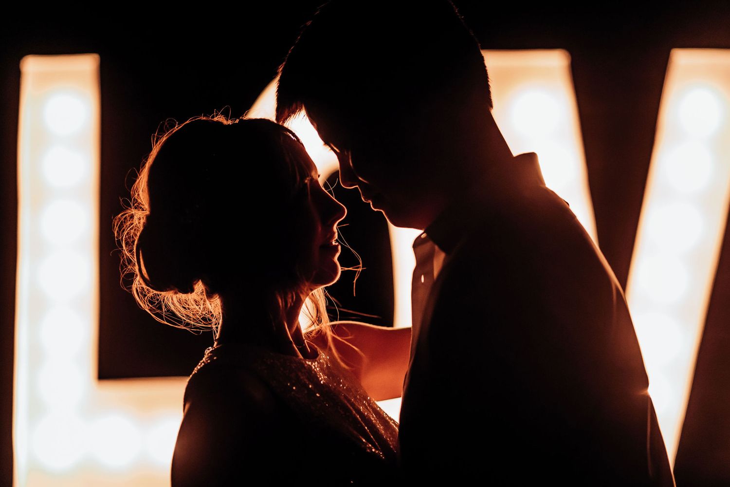 Silhouetted couple sharing intimate moment in front of illuminated decor at wedding reception.