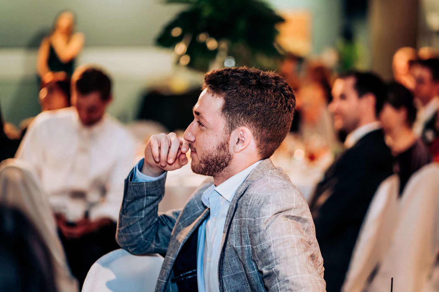 A thoughtful guest in a grey suit jacket listens intently during an indoor event.