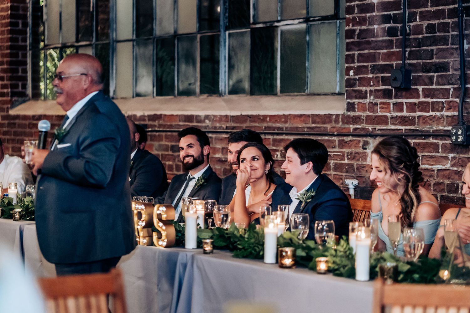 Reception speaker addresses wedding guests seated at decorated tables in brick-walled venue.