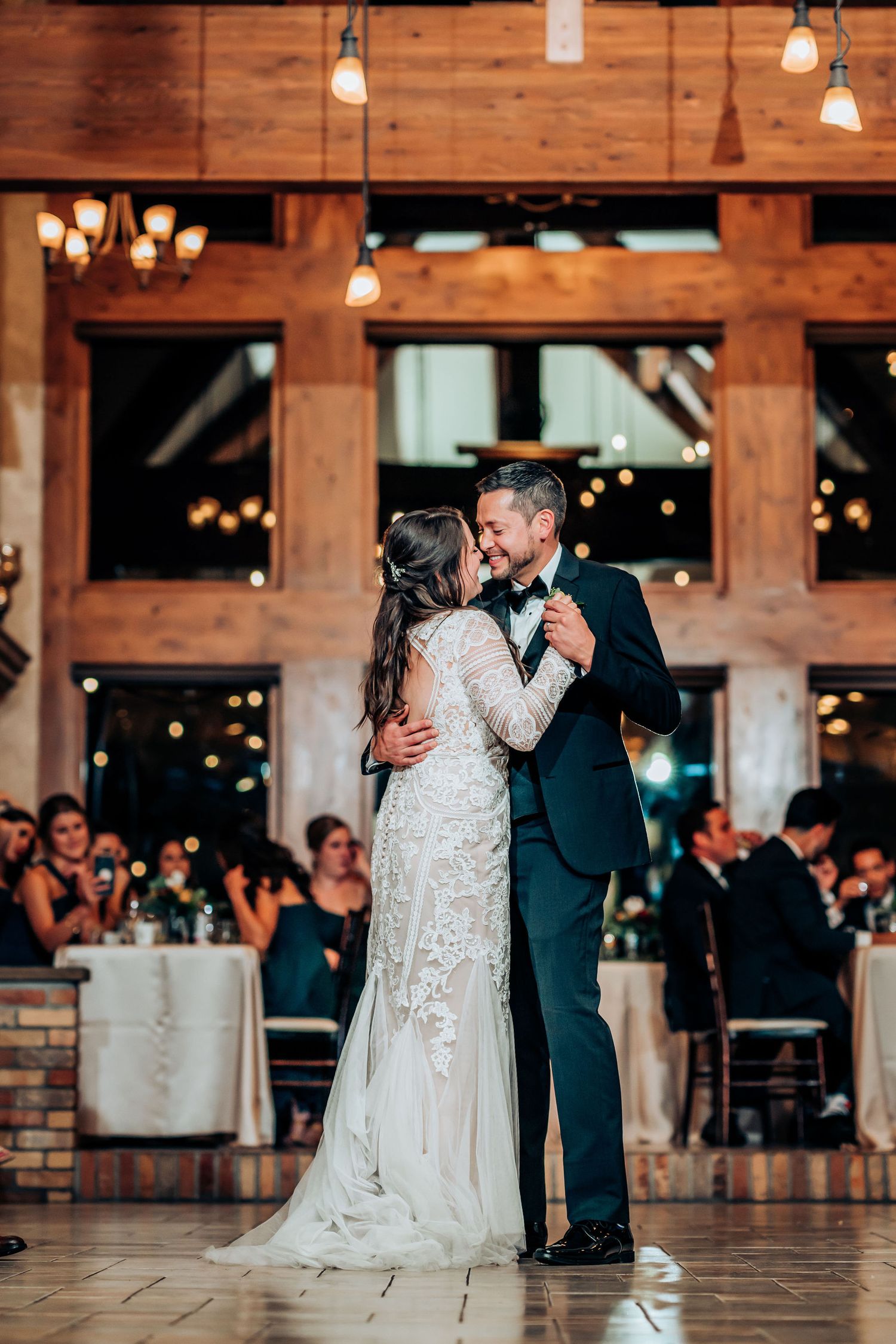 Couple shares a romantic dance in an elegant rustic venue with wooden details and warm lighting.