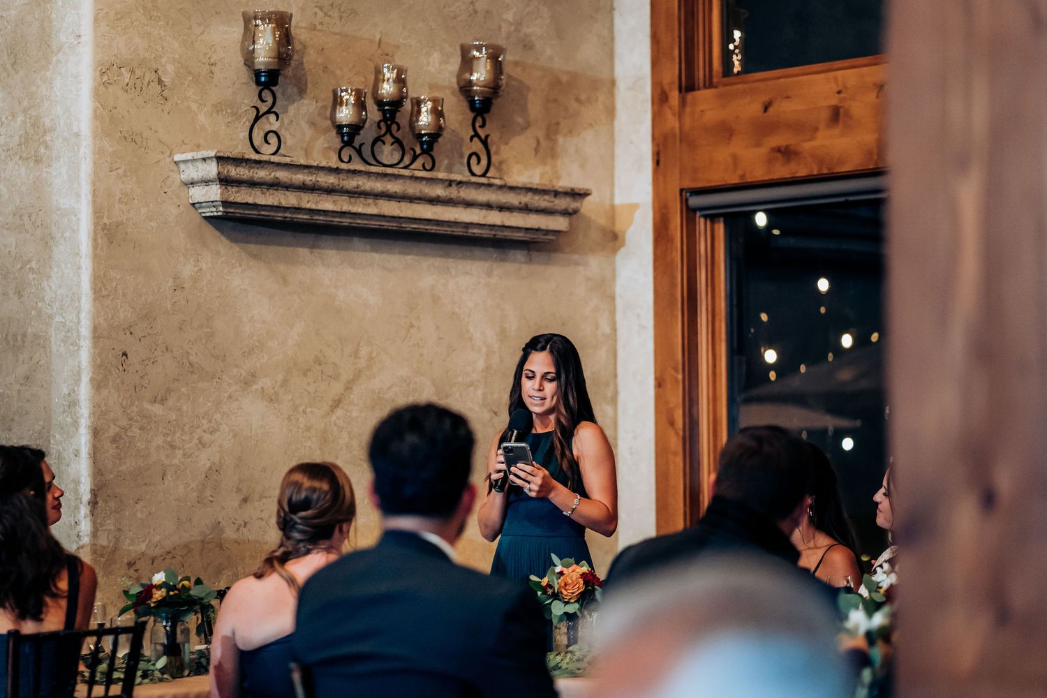 A speaker addresses wedding guests during a reception in a rustic venue with stone walls.