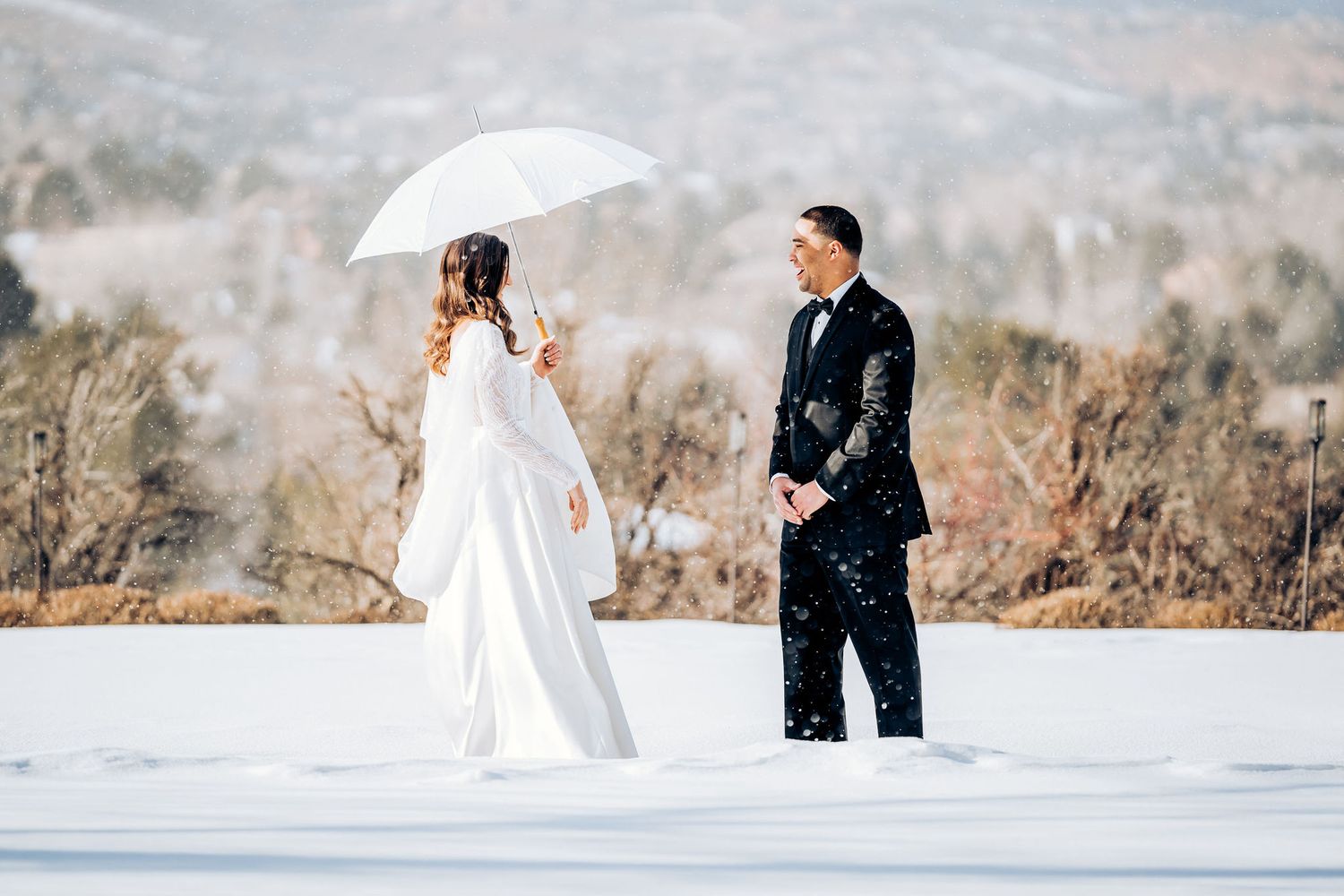 A couple shares a moment in the snow with a white umbrella and mountain scenery.