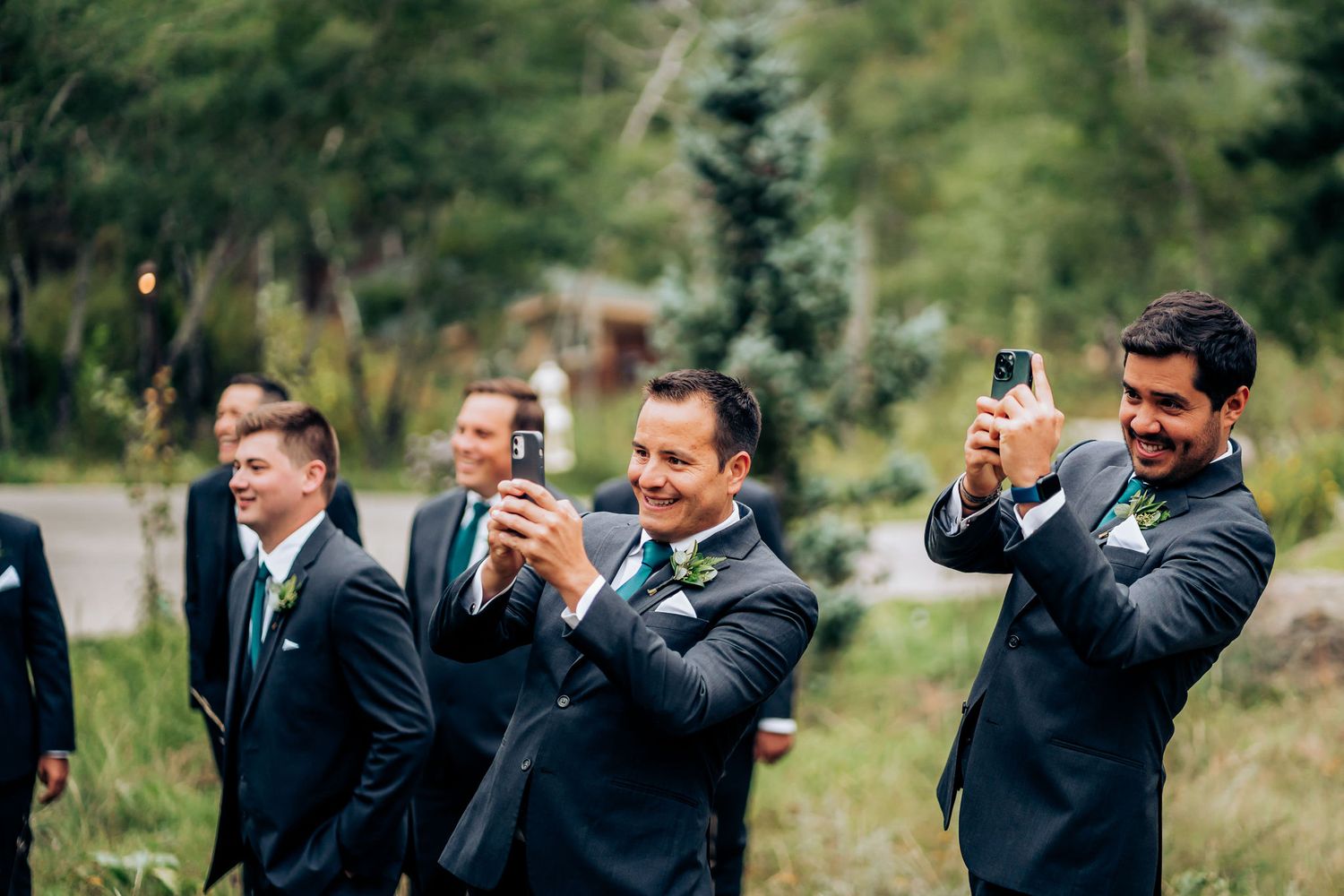 Groomsmen in dark suits take photos on their phones during outdoor wedding celebration.