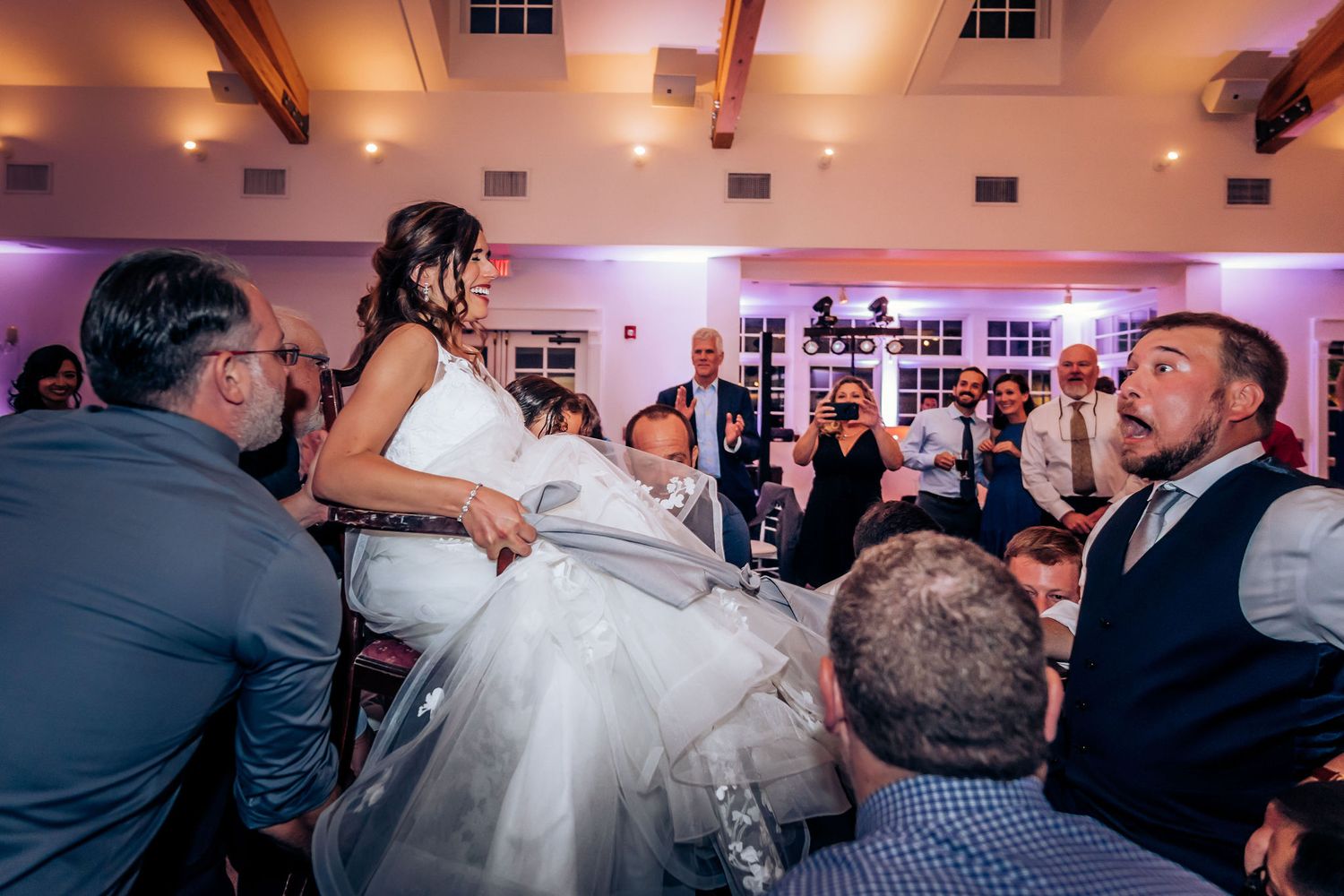 Wedding guests lift the bride seated in a chair during traditional reception celebrations.