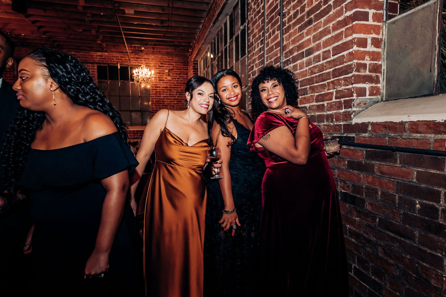 Wedding party poses in elegant dresses against exposed brick wall with warm lighting.