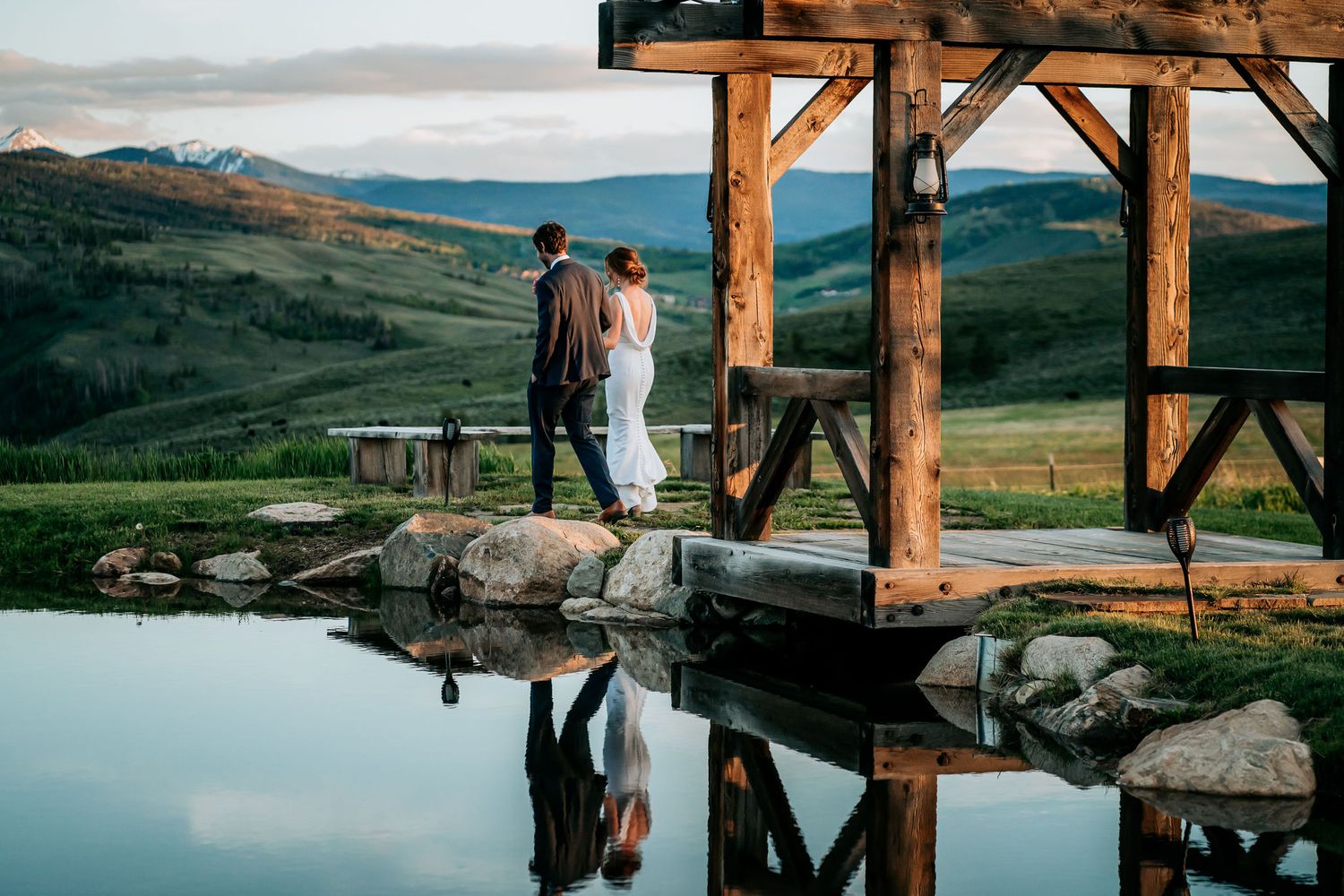 A silhouetted couple stands on a rustic wooden deck with mountain views and reflective pool.