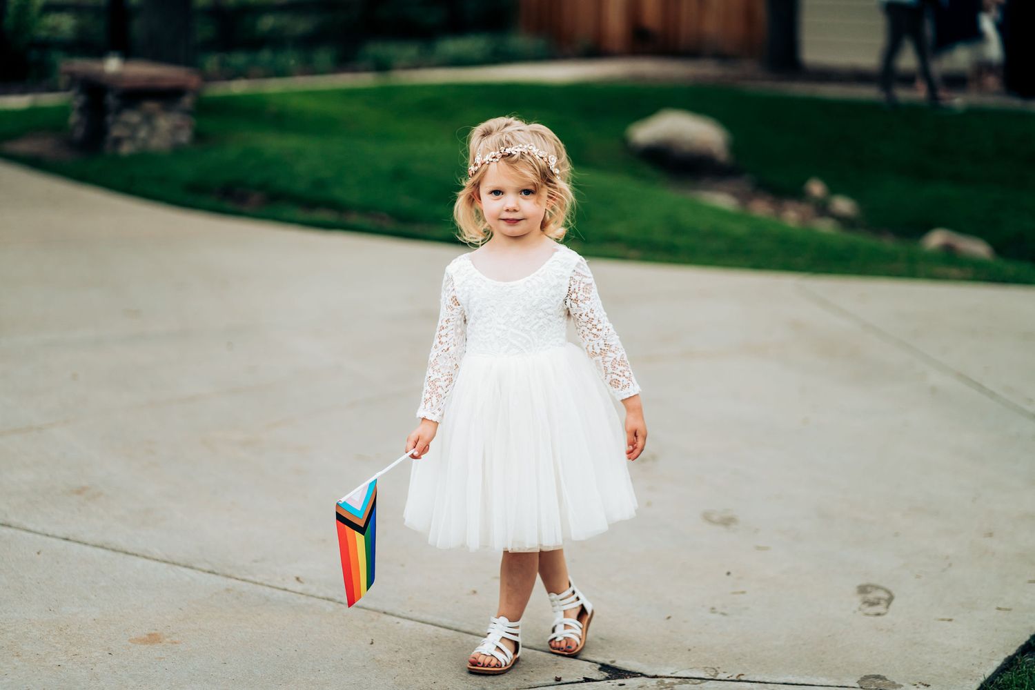 Small child in white lace dress holding colorful book on outdoor walkway.