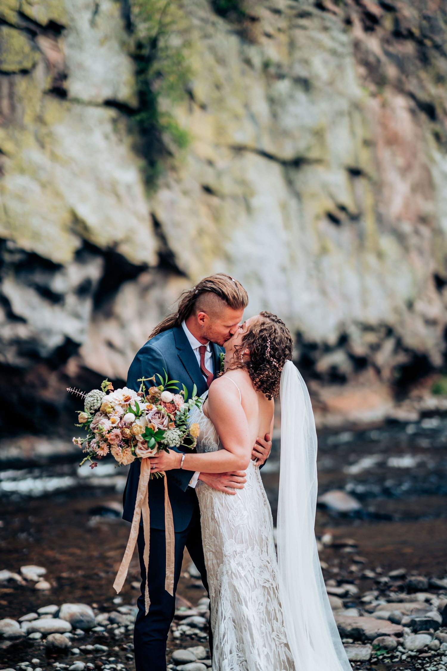 Intimate wedding moment captured against dramatic cliffside with flowing river and natural light.