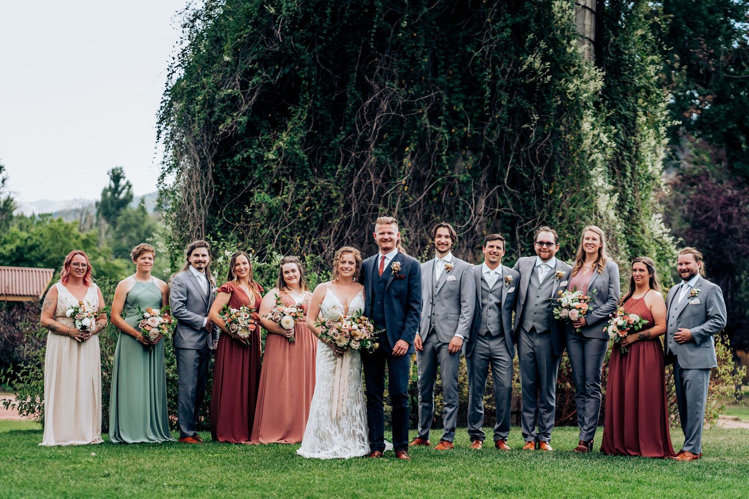 Large wedding party with bridesmaids in mix-matched dresses poses in front of rustic venue.