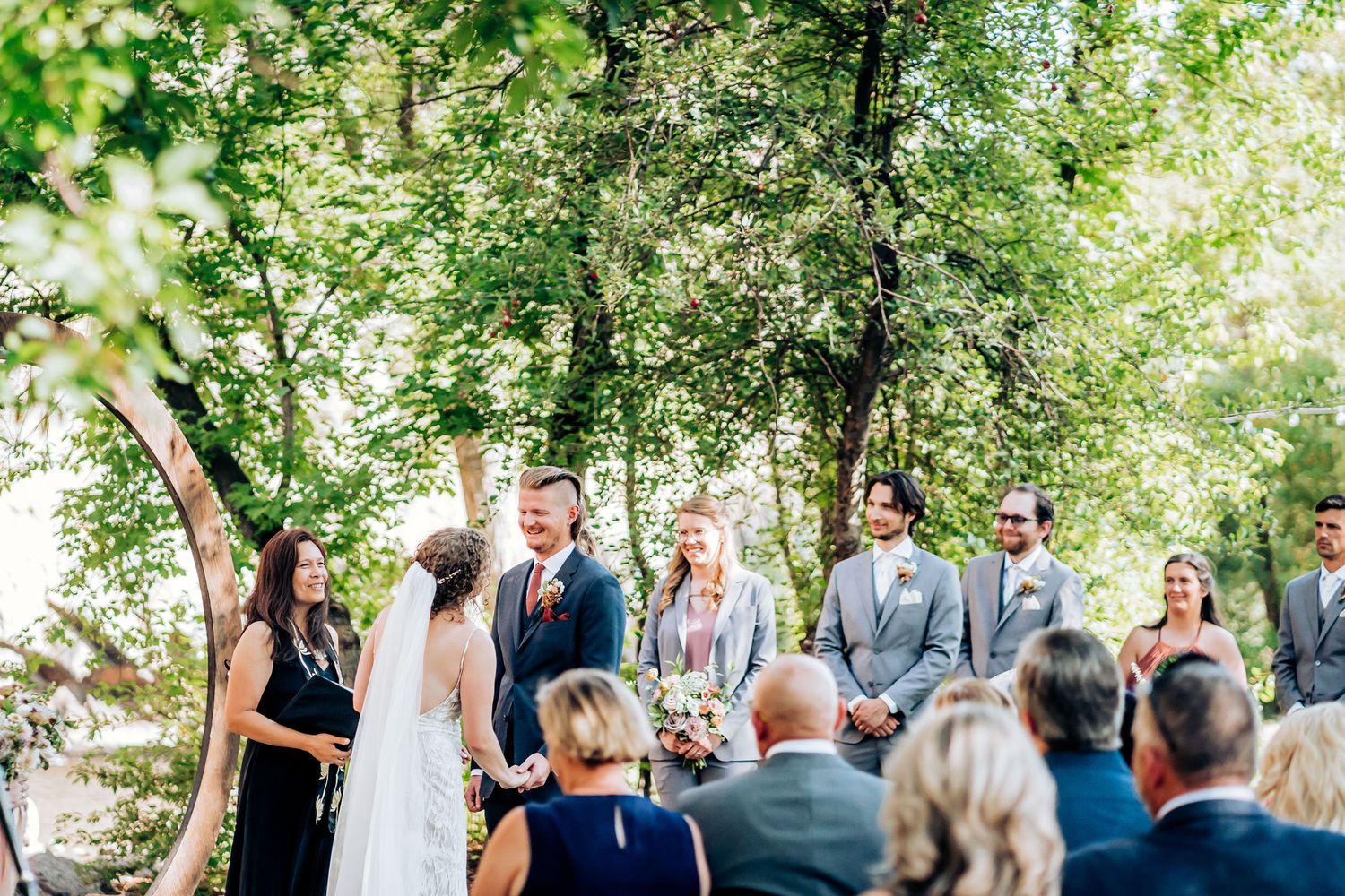 Wedding ceremony in progress under tree canopy with gathered guests and wedding party.