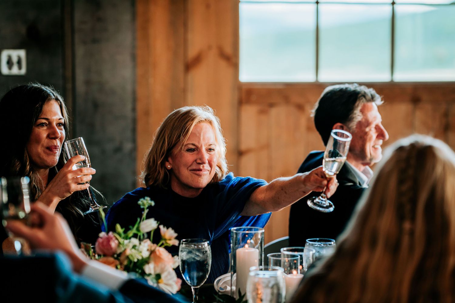Guests raise their glasses in a toast during an indoor celebration with floral centerpieces and natural window lighting.