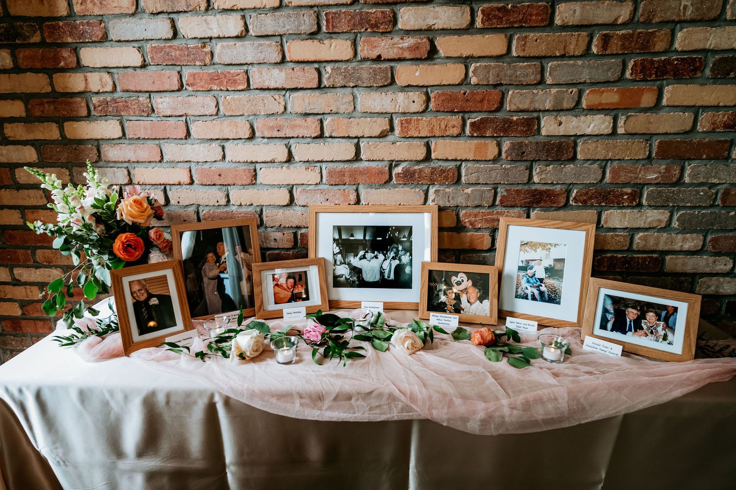 A memorial table display with framed photos and floral arrangements against a rustic brick wall.