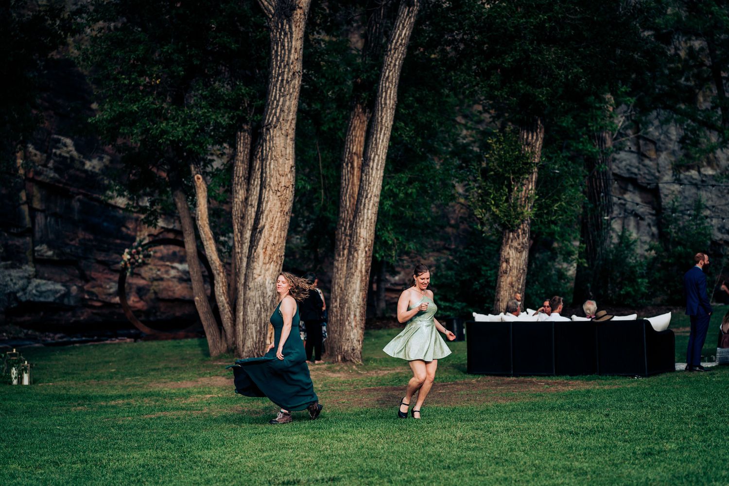 Two people dance playfully on a grassy lawn with tall trees and a buffet table in the background.