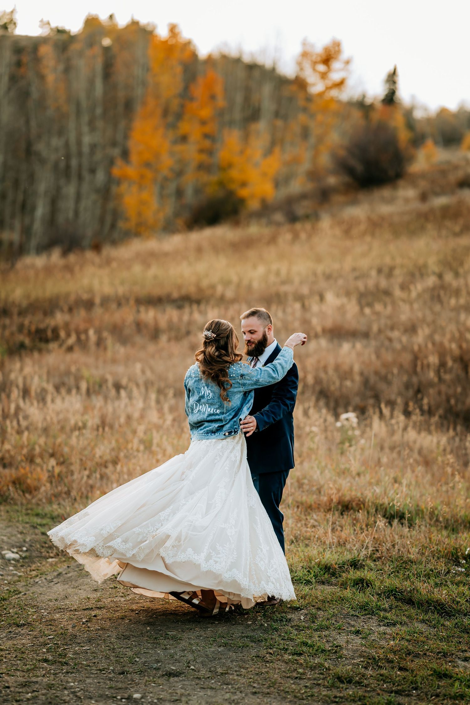 A couple dances in a field with golden autumn trees in the background.