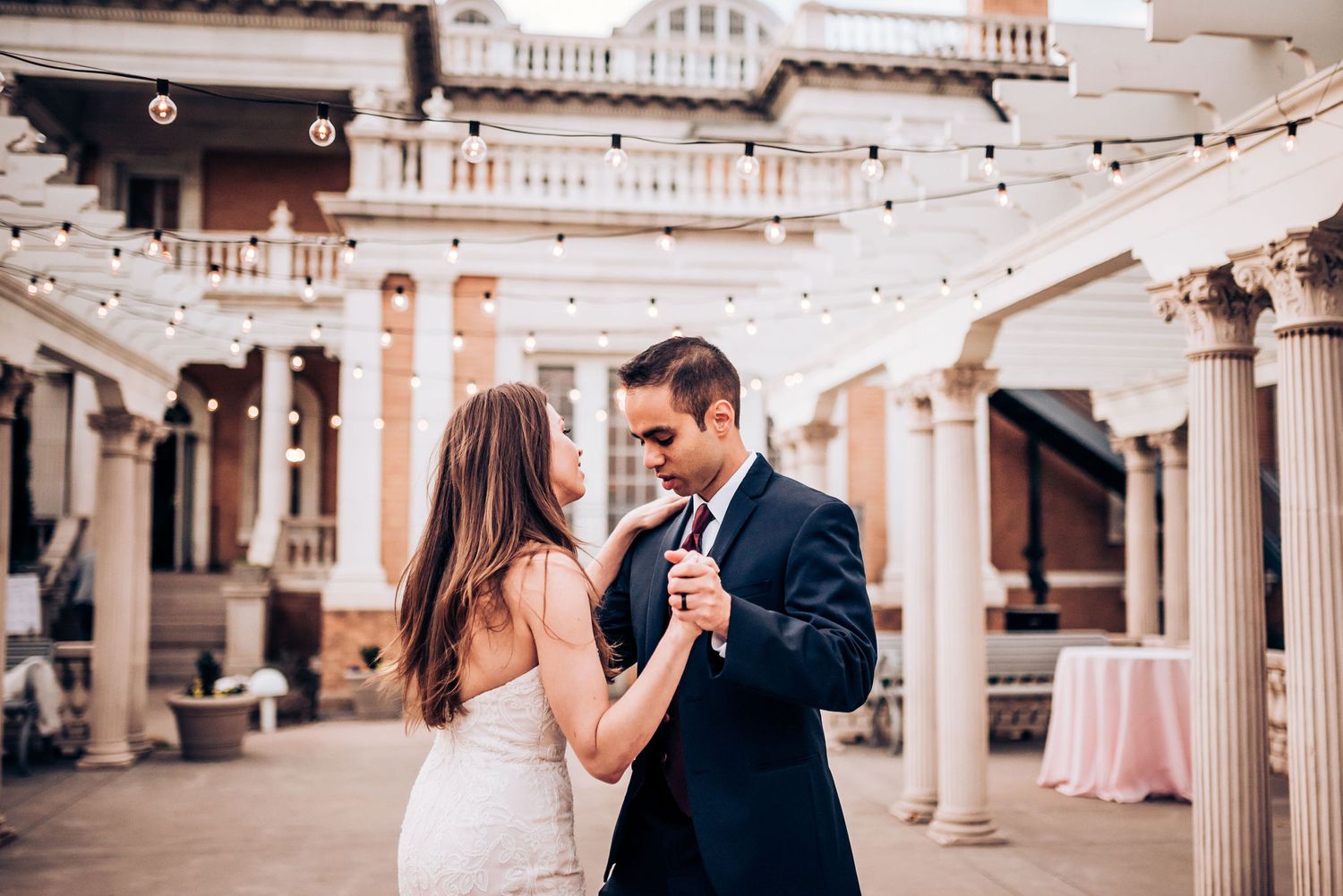 Newlyweds dancing in elegant ballroom with classical columns and string lights overhead.