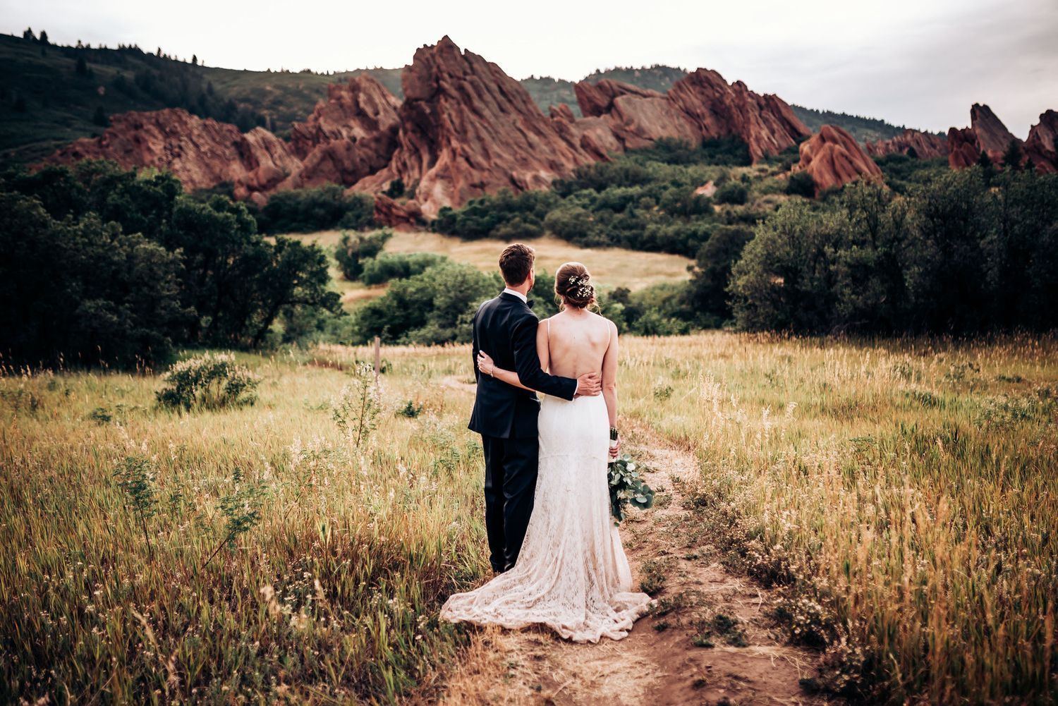A wedding couple admires the view of red rock formations while standing on a mountain trail.