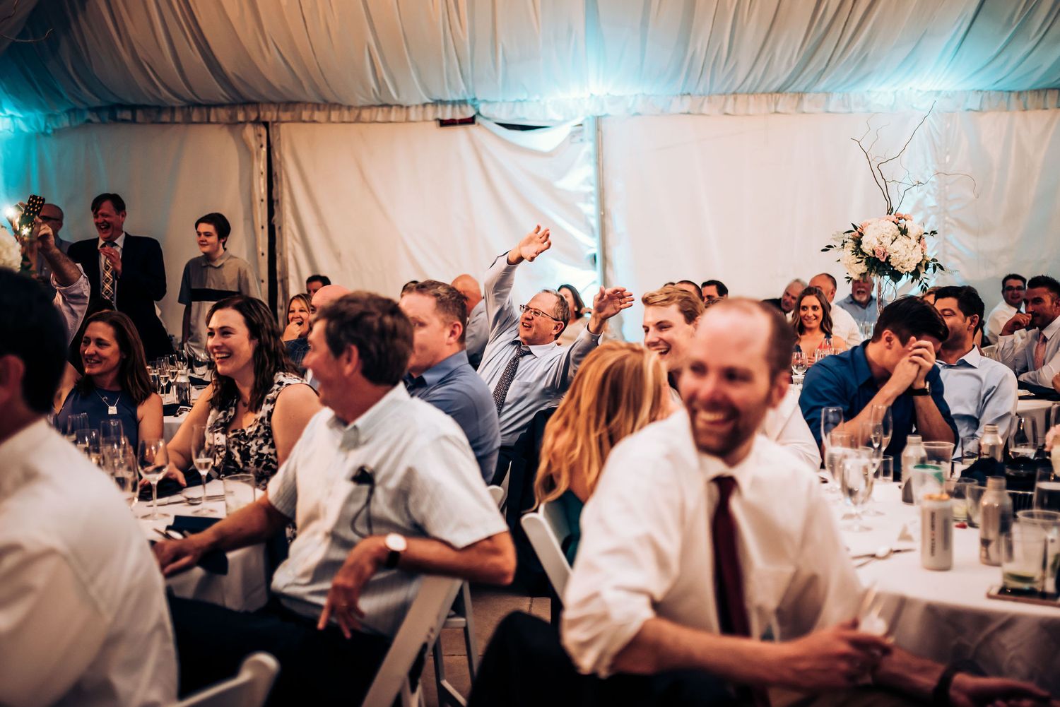 Wedding reception guests cheering and celebrating in white tent venue.
