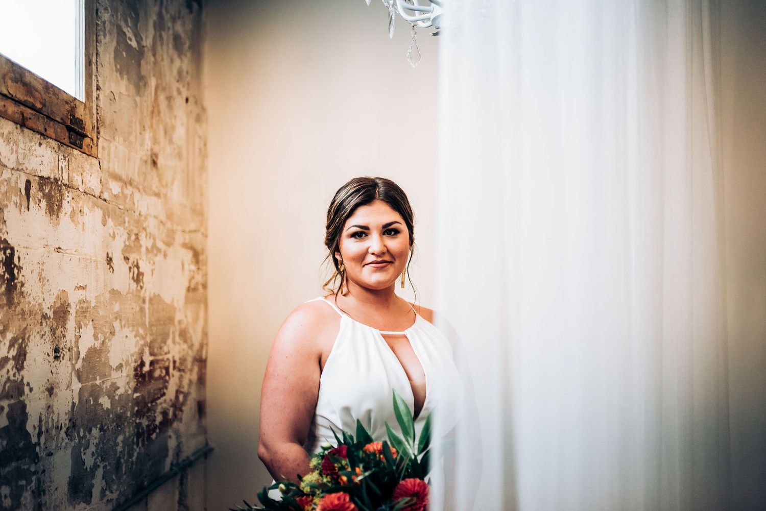 Bridal portrait in white gown against textured wall holding vibrant bouquet.