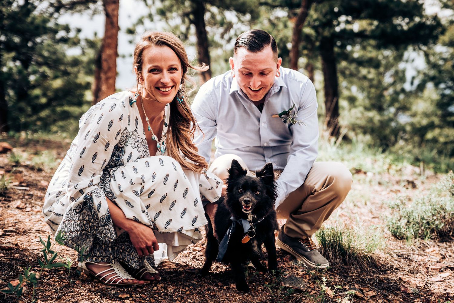 A couple poses outdoors with their black dog among pine trees on a sunny day.
